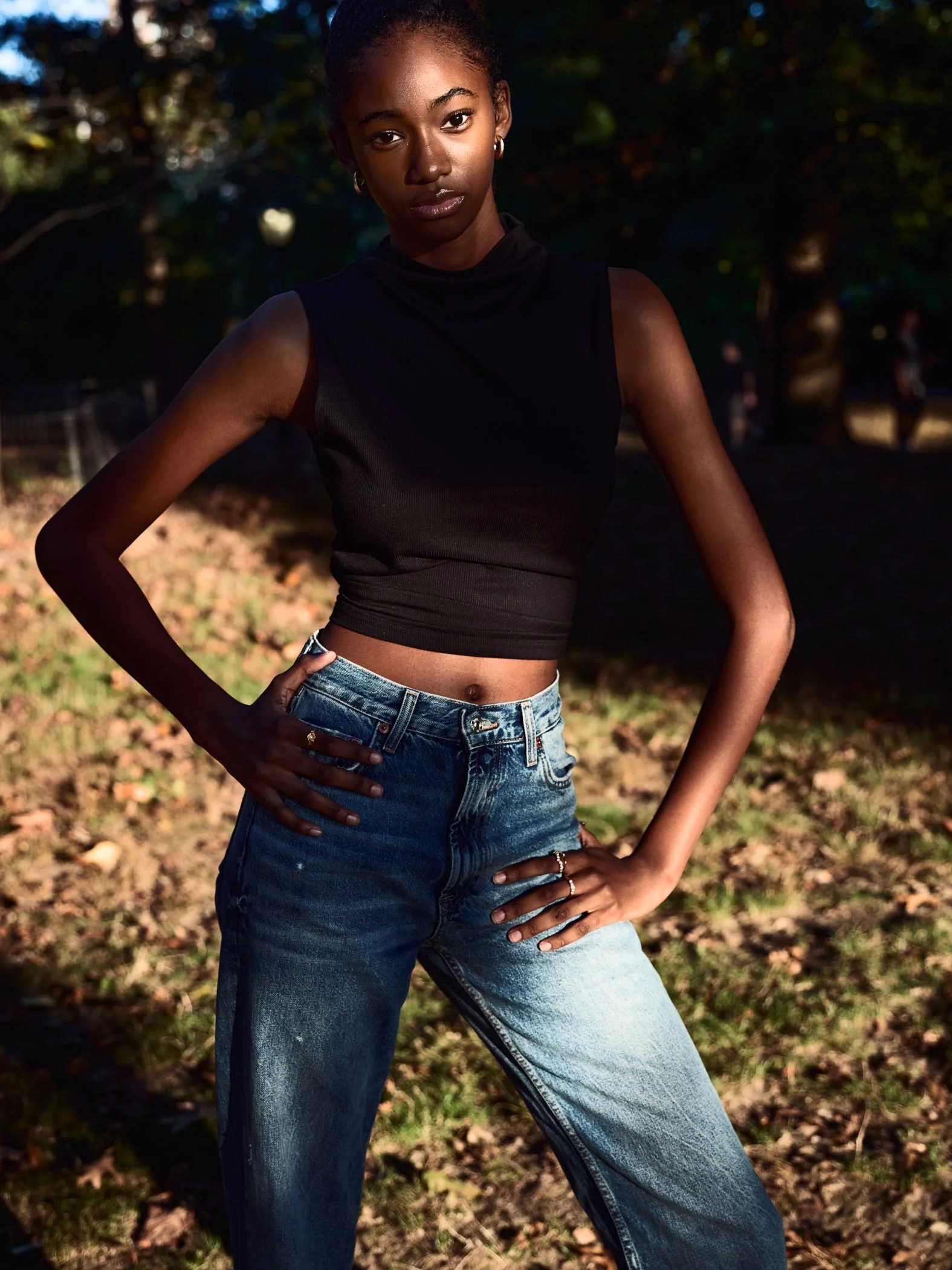 A young woman with dark skin and short hair posing outdoors in a black sleeveless top and high-waisted blue jeans with her hands on her hips, standing in a park during the daytime.