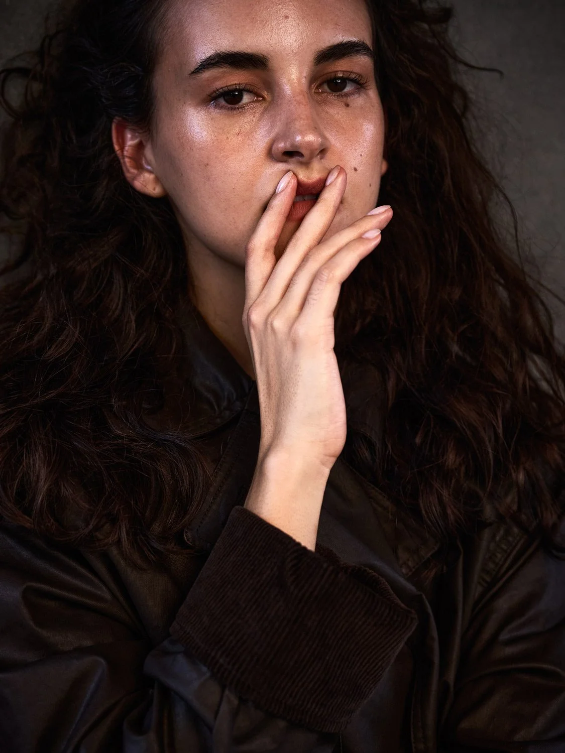 A woman with curly brown hair and fair skin sits with her hand near her face, wearing a dark brown leather jacket.