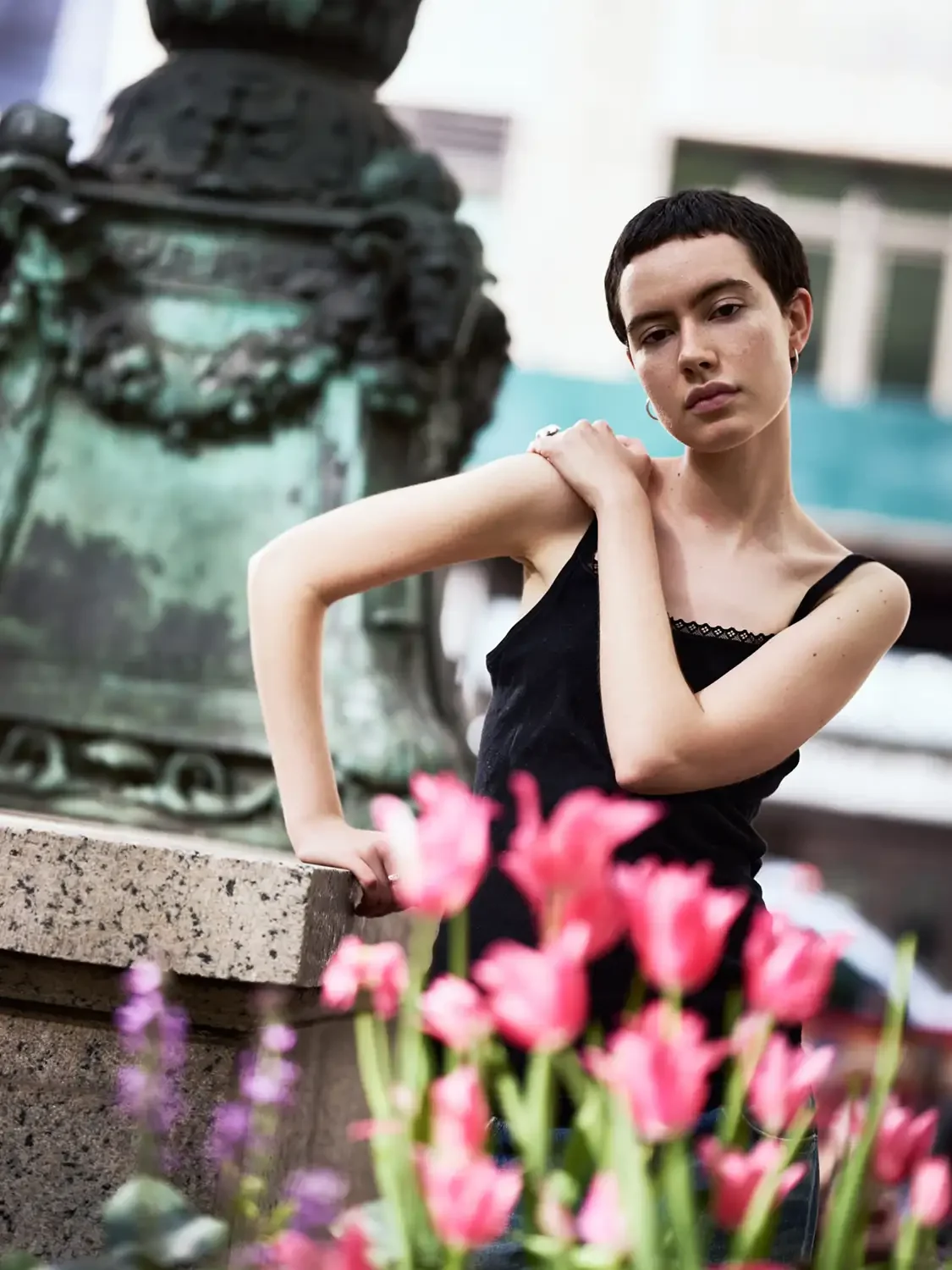 A young woman with short dark hair and pale skin is posing outdoors next to a flower bed with pink and purple flowers. She is wearing a black sleeveless top and has one hand resting on her shoulder. In the background, an abstract sculpture is visible