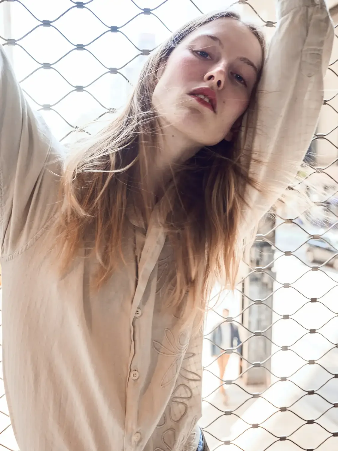 A young woman with long hair leaning against a chain-link fence, looking at the camera with a relaxed expression.