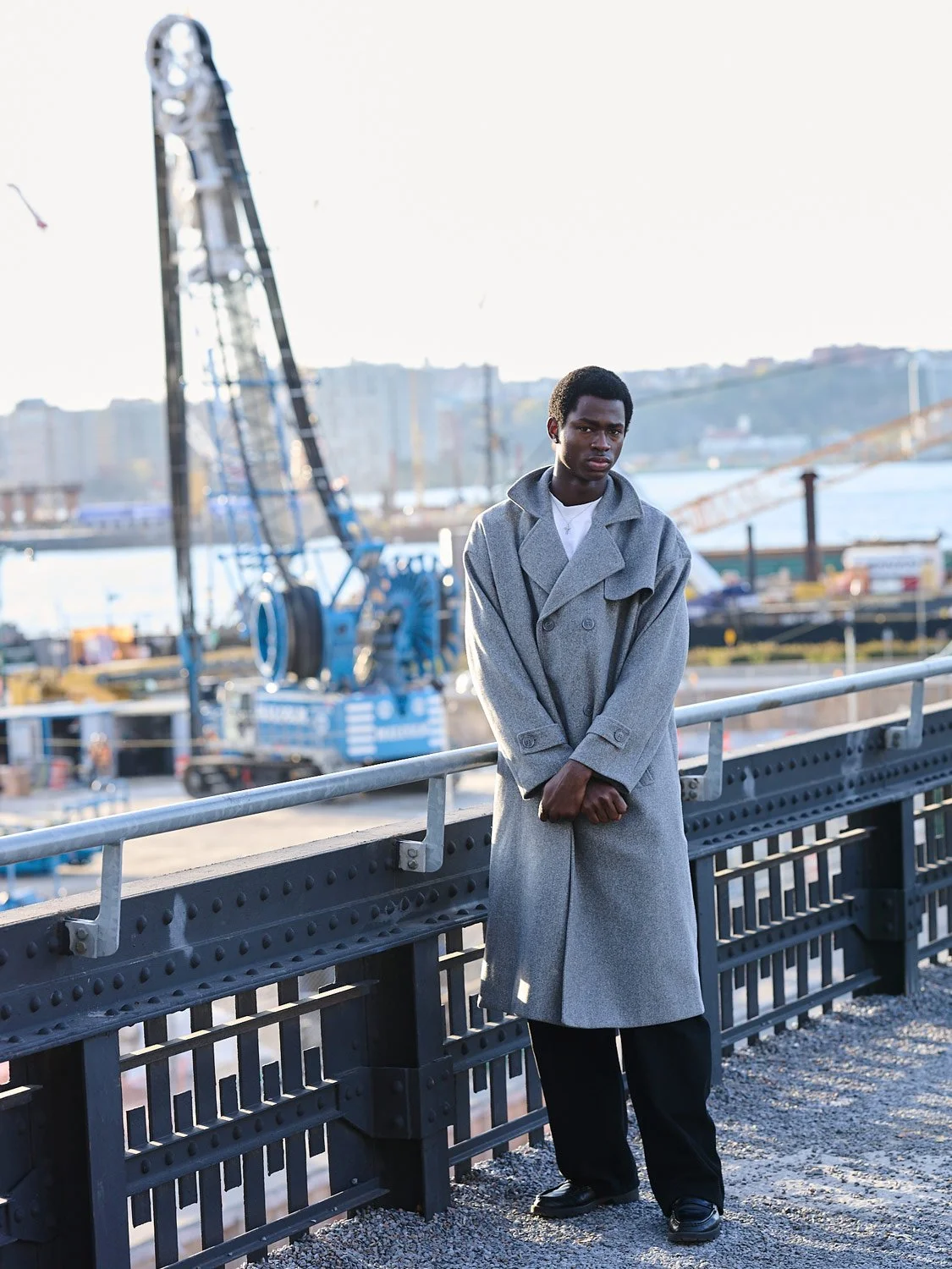 A young man wearing a gray coat and black shoes leaning on a railing at a waterfront area with cranes and industrial structures in the background during daylight.