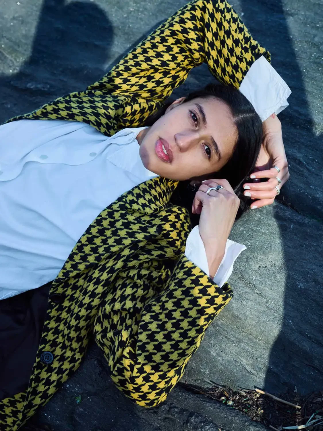 A woman lying on her back on a concrete surface, looking at the camera with her arms behind her head. She is wearing a yellow and black houndstooth patterned jacket, a white shirt, and multiple rings on her fingers.