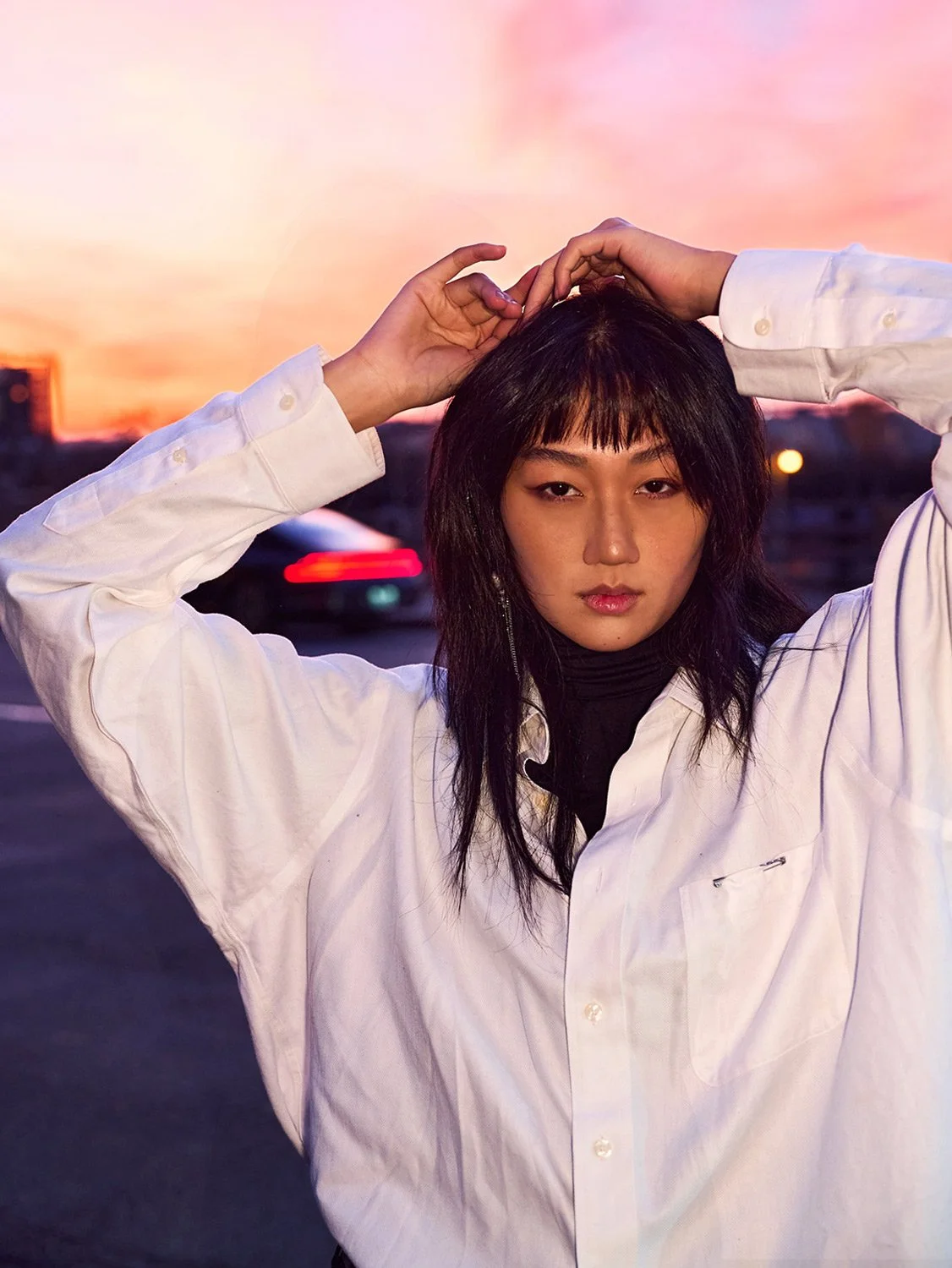 Young woman with black hair and bangs, wearing a white button-up shirt over a black turtleneck, posing outdoors during sunset with a pink and orange sky in the background.