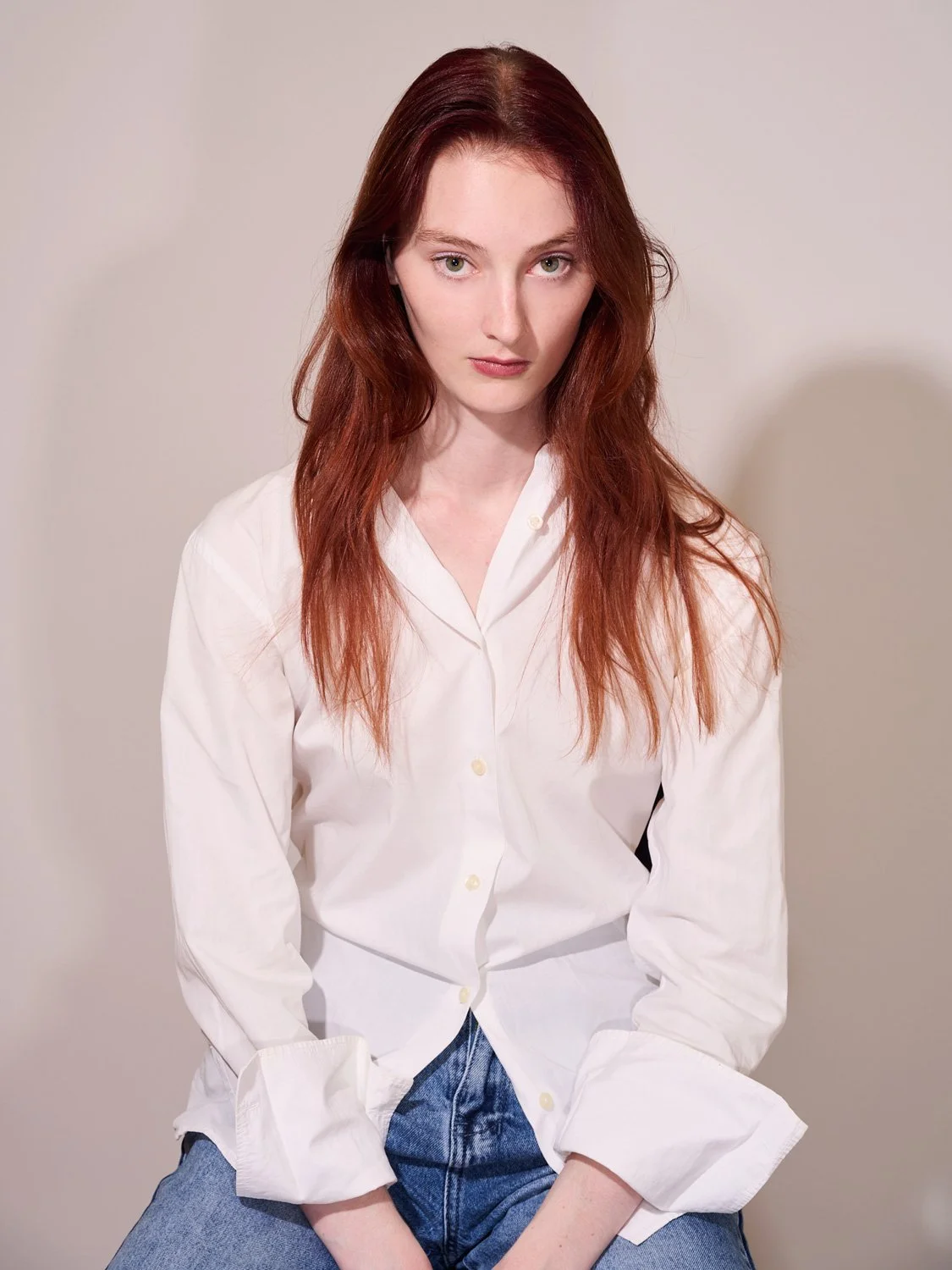 A young woman with long red hair, wearing a white button-up shirt and blue jeans, sitting against a plain light-colored background.