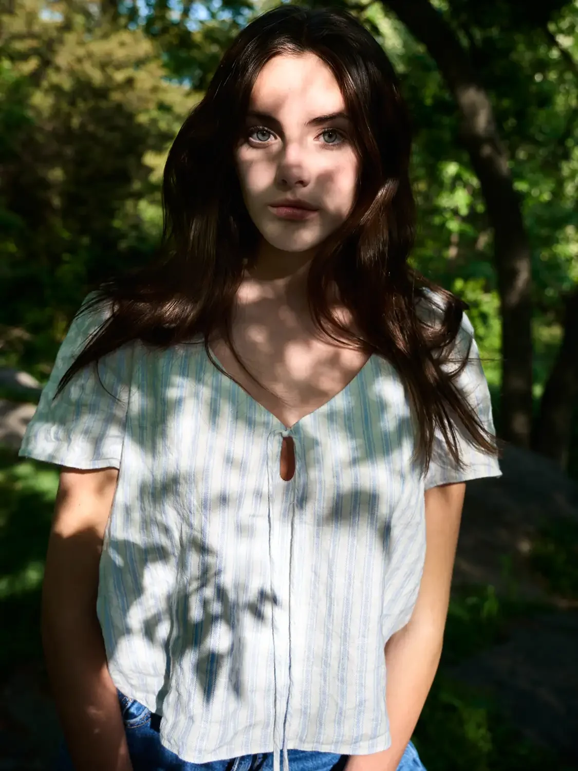 Young woman with brown hair and blue eyes standing outdoors in a forest with sunlight filtering through the trees.