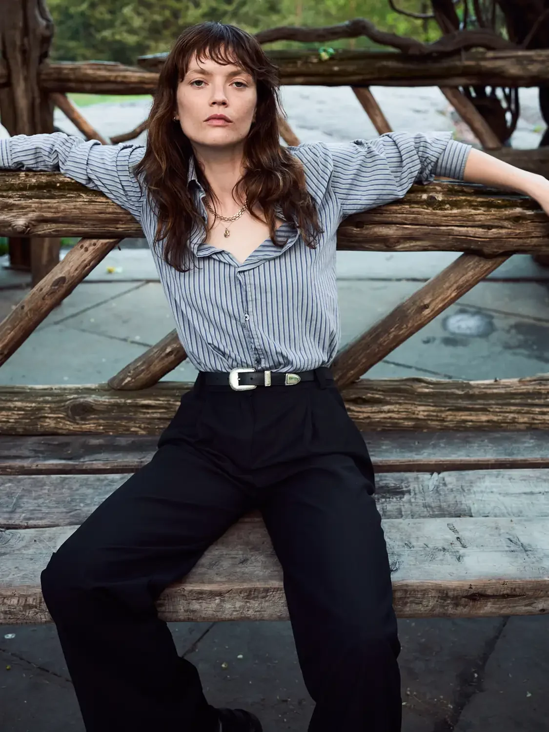A woman with curly brown hair wearing a striped button-up shirt and black pants, sitting on a wooden bench with a rustic wooden fence behind her, outdoors.