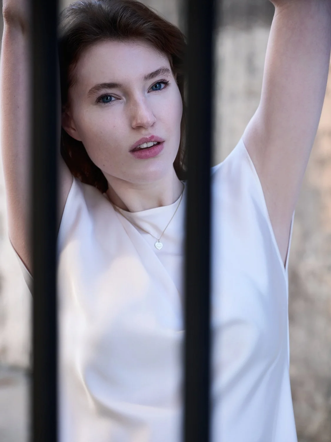 A young woman with reddish brown hair and blue eyes is looking directly at the camera with her arms raised, framing her face, in an outdoor setting with blurred background.