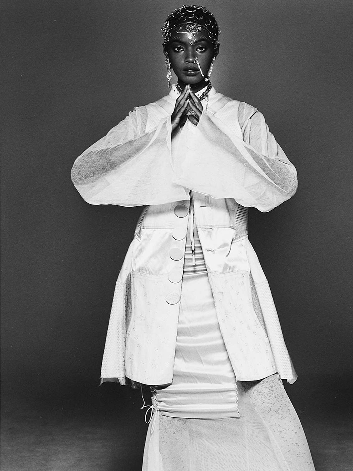 Black and white photo of a woman wearing elaborate jewelry and layered clothing, with her hands pressed together in front of her chest.