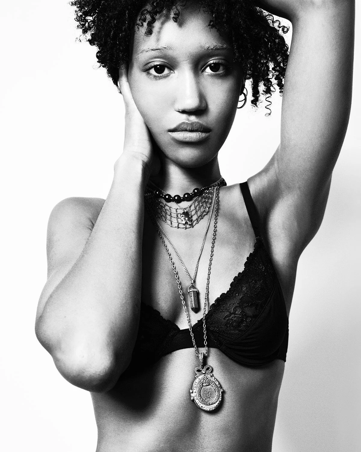 Black and white portrait of a young woman with curly hair wearing layered necklaces, a lace bra, and jewelry, looking directly at the camera with one hand resting on her face and the other arm raised above her head.