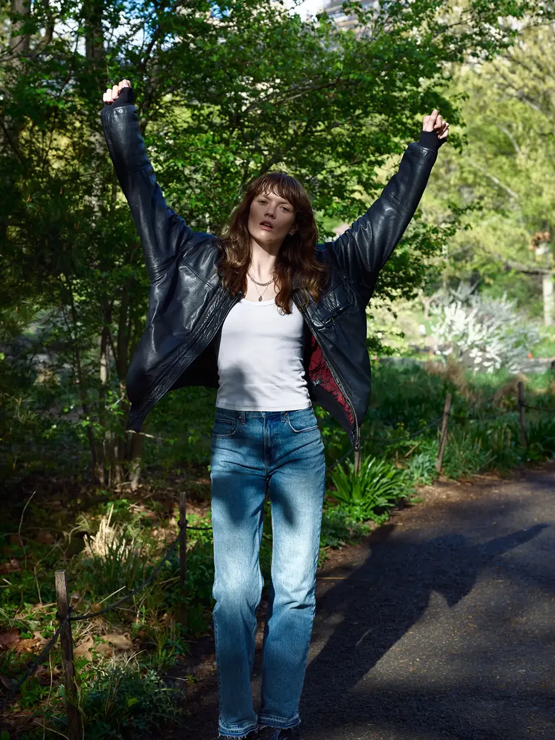 A woman with brown hair wearing a black leather jacket, white shirt, and blue jeans, standing outdoors on a pathway with greenery and trees in the background, arms raised in a triumphant pose.