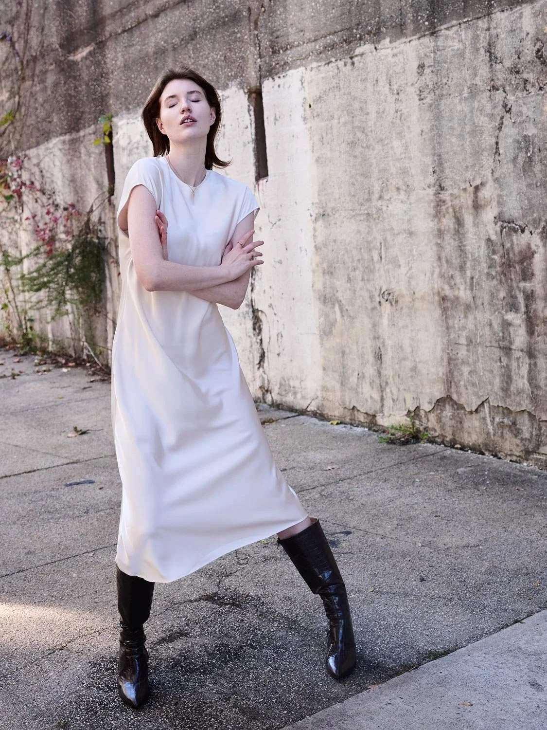 A woman with dark hair wearing a white dress and black boots stands with her arms crossed against a weathered concrete wall outdoors.