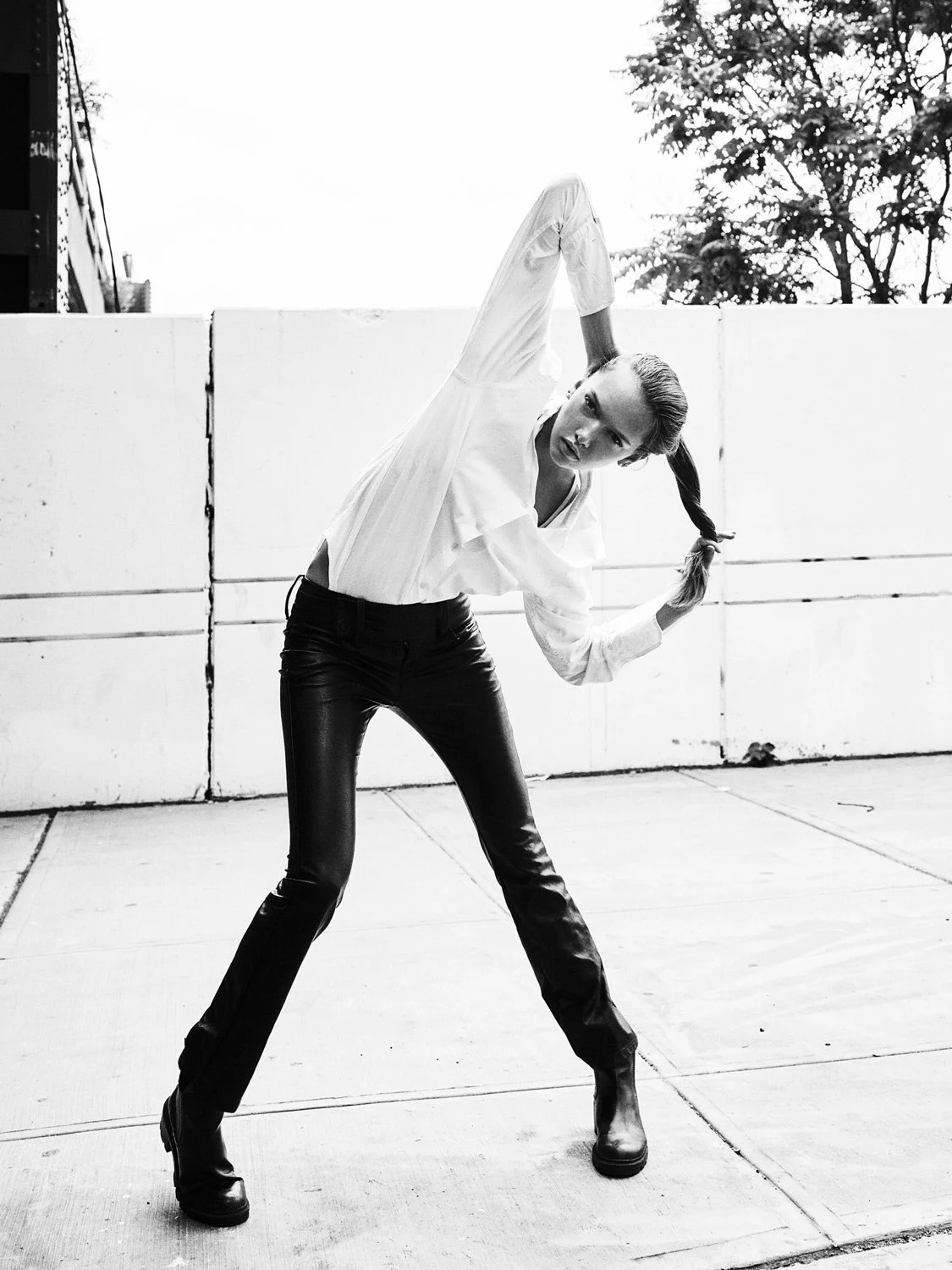 A woman in a white shirt and black leather pants poses outdoors, holding her braided hair and leaning forward against a white fence, with trees and sky in the background.