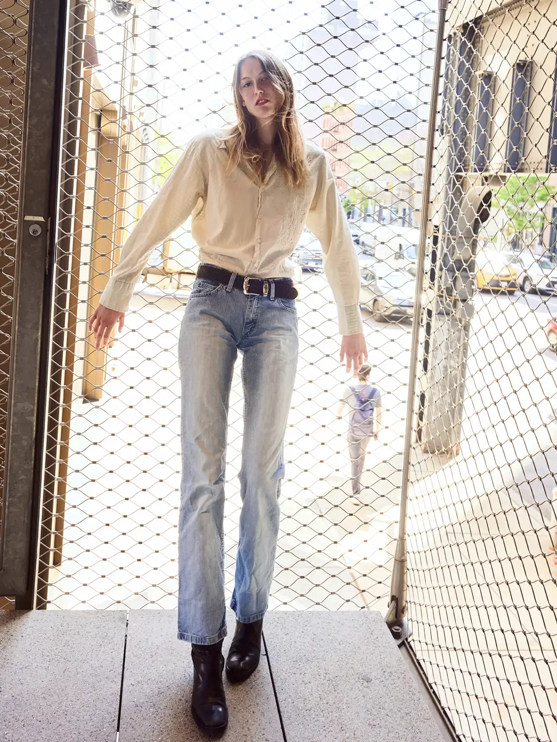 Woman standing behind a metal gate on a city sidewalk, wearing a fashionable  light-colored shirt, jeans, and black boots.