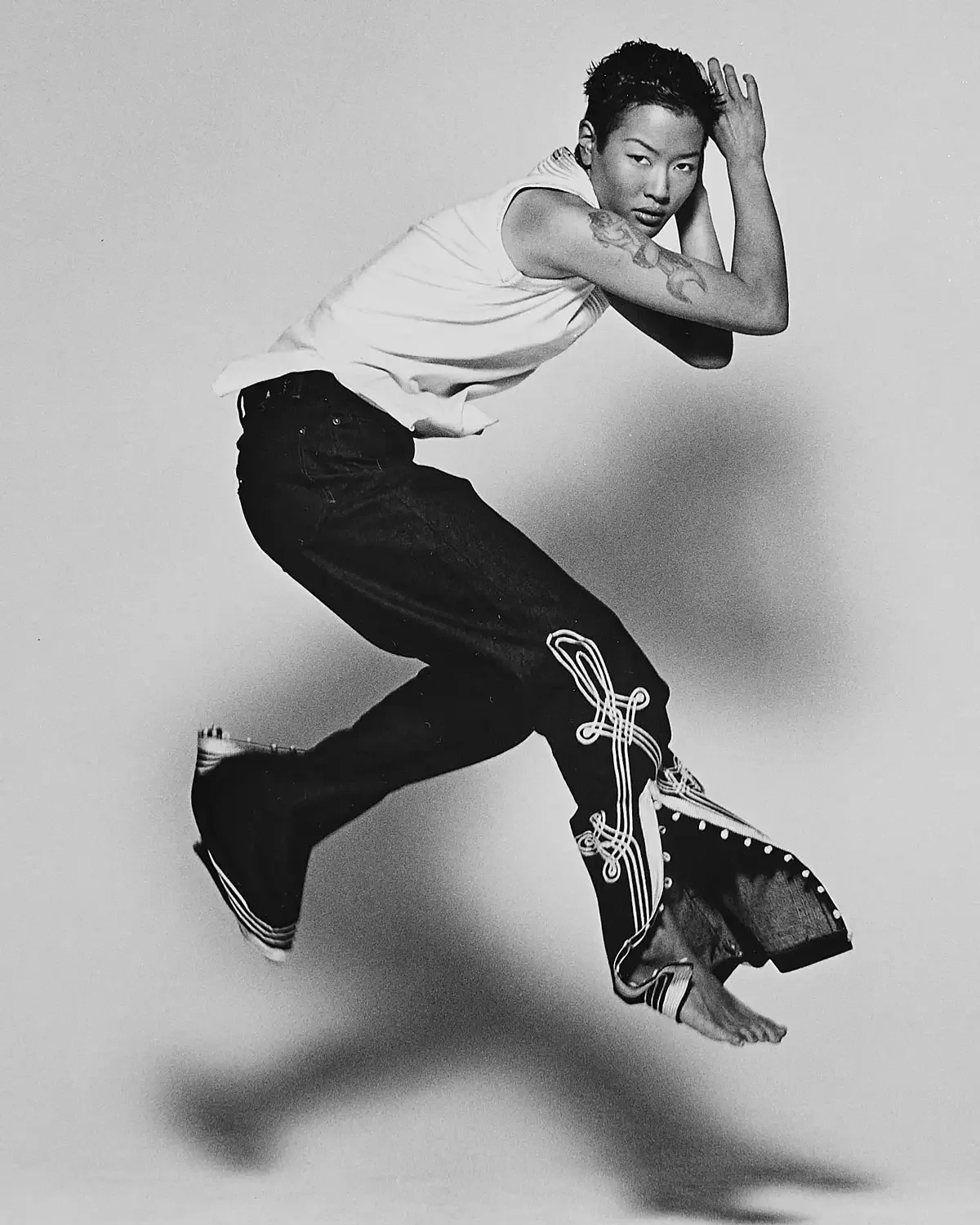 Jenny Shimizu mid-air in a dance pose, wearing a djellaba top and jeans with tattoos on her arm, for a fashion shooting in a photo studio black and white picture.