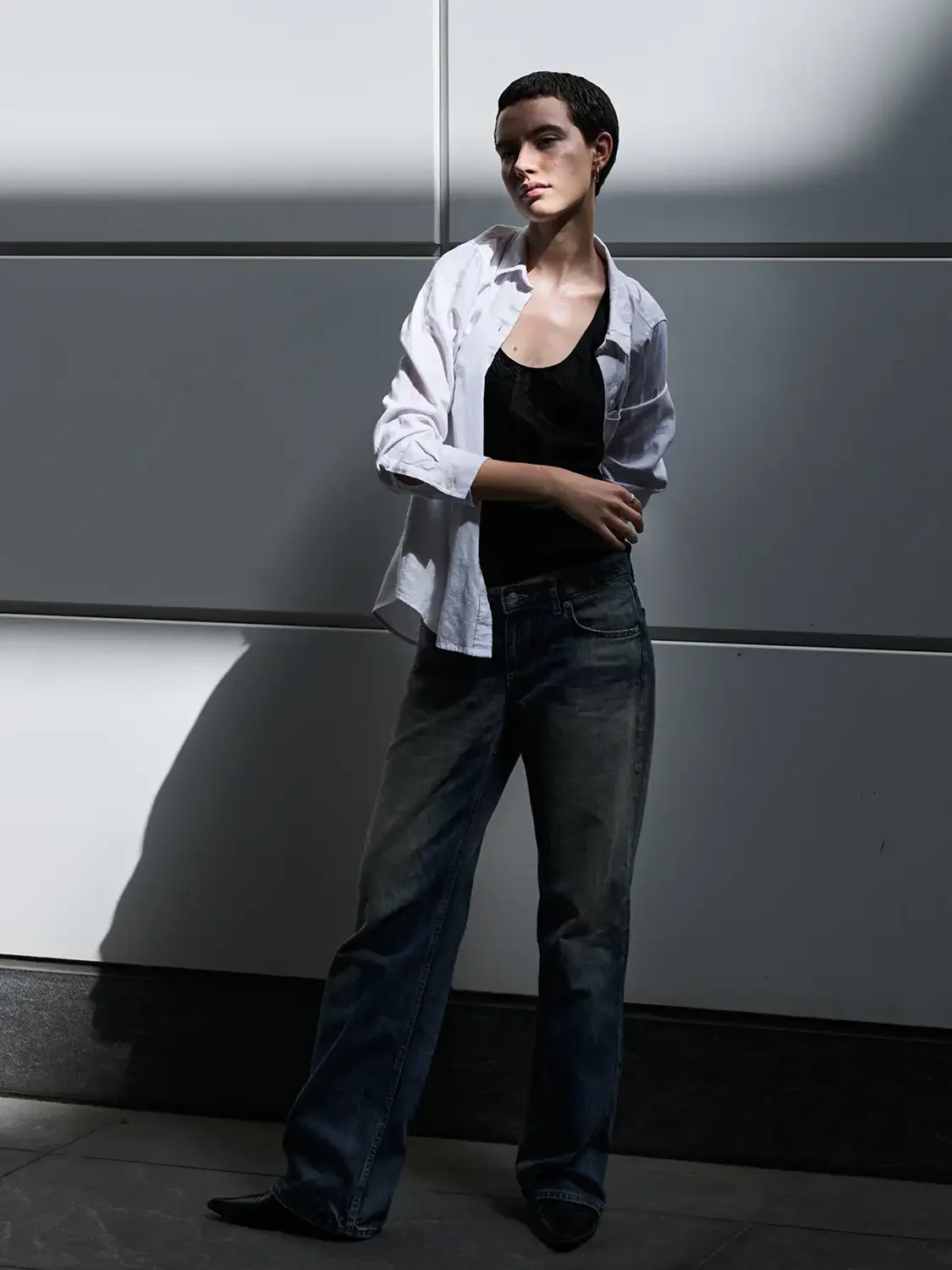 A woman with short dark hair standing against a gray wall, wearing a white button-up shirt over a black top and loose-fitting dark jeans.