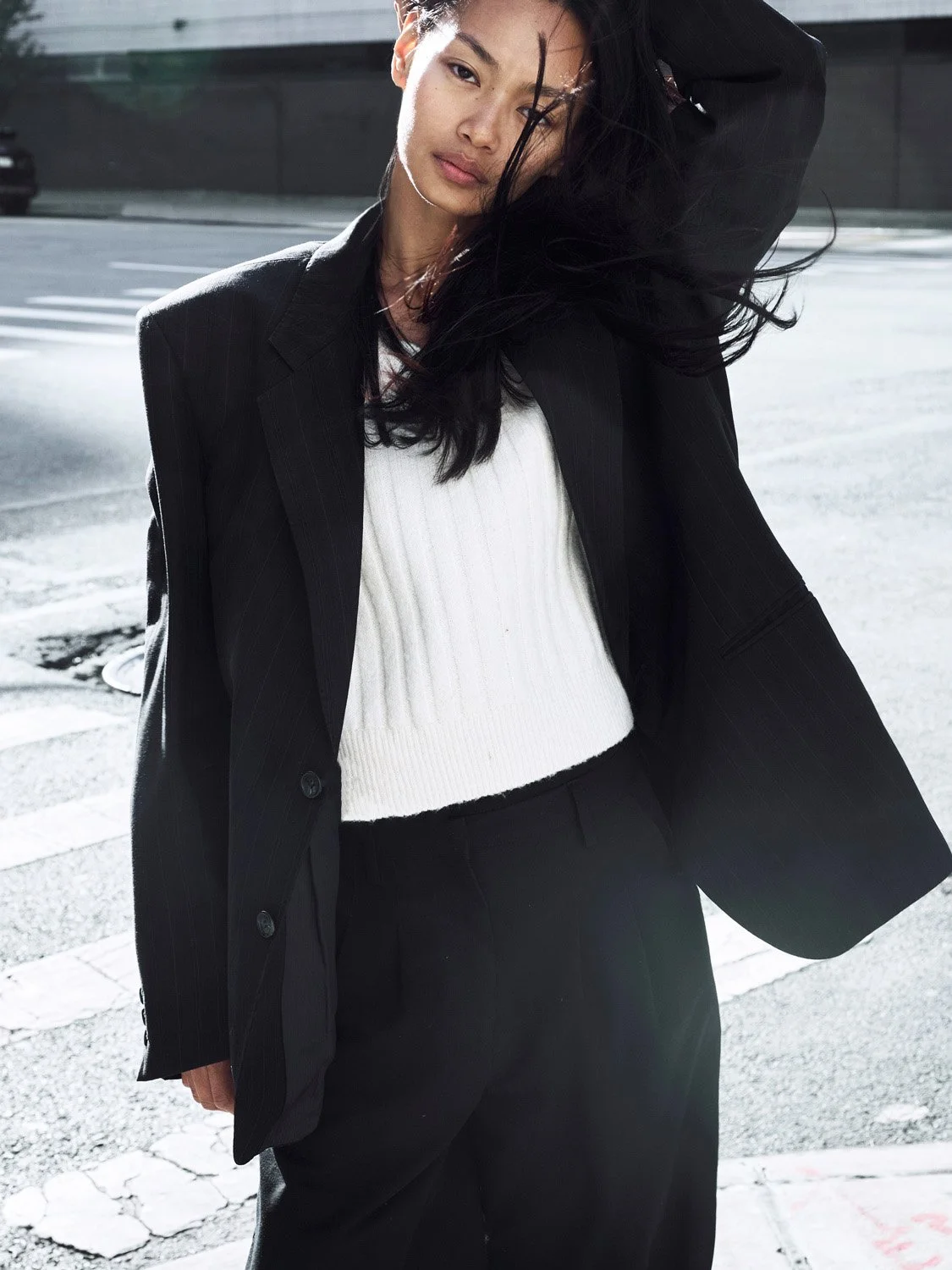 A woman standing on a city street, wearing a black blazer, white sweater, and black pants, with wind blowing her hair.