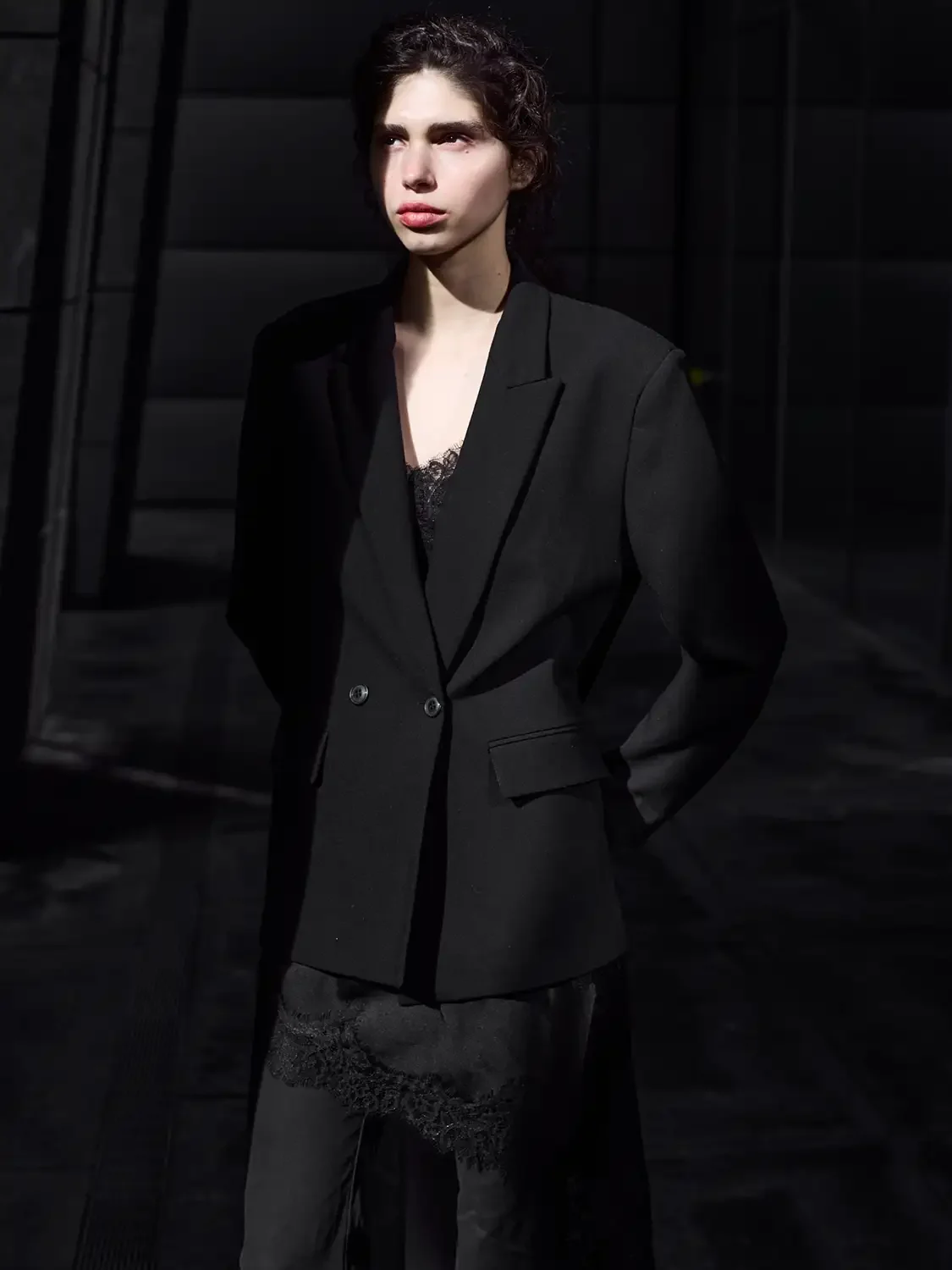 A woman with dark, curly hair wearing a black blazer over a lacy top standing in a dark, modern setting.