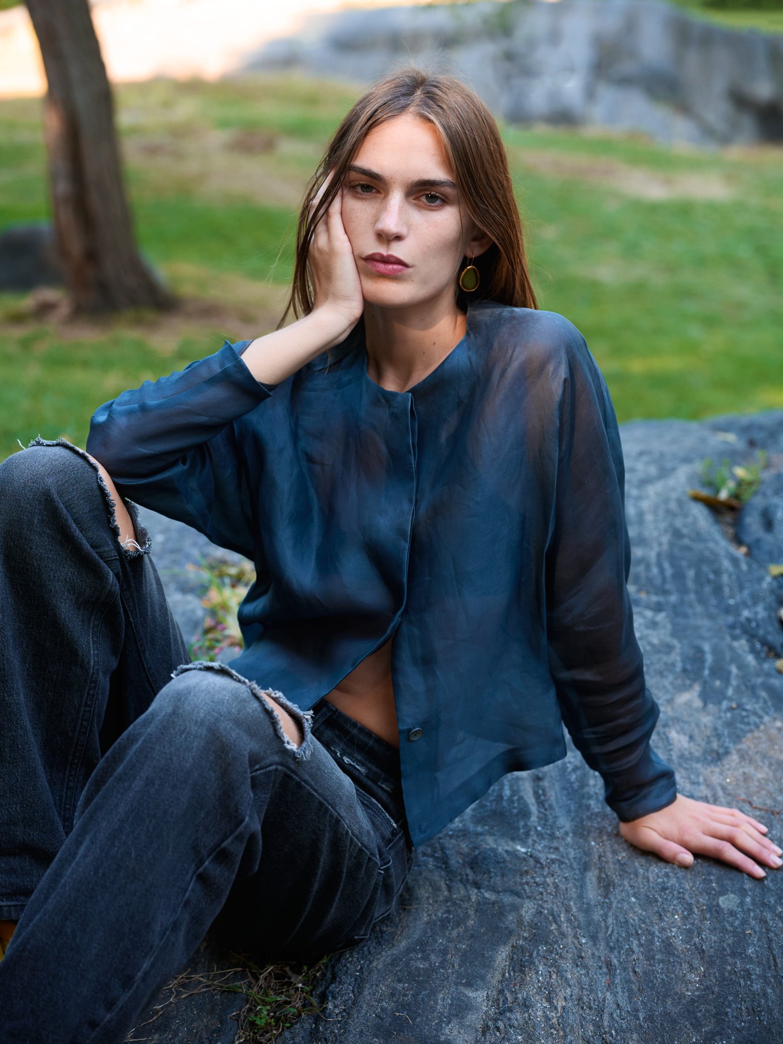 A young woman with long brown hair, sitting on a large rock outdoors, wearing a sheer blue blouse and ripped black jeans.