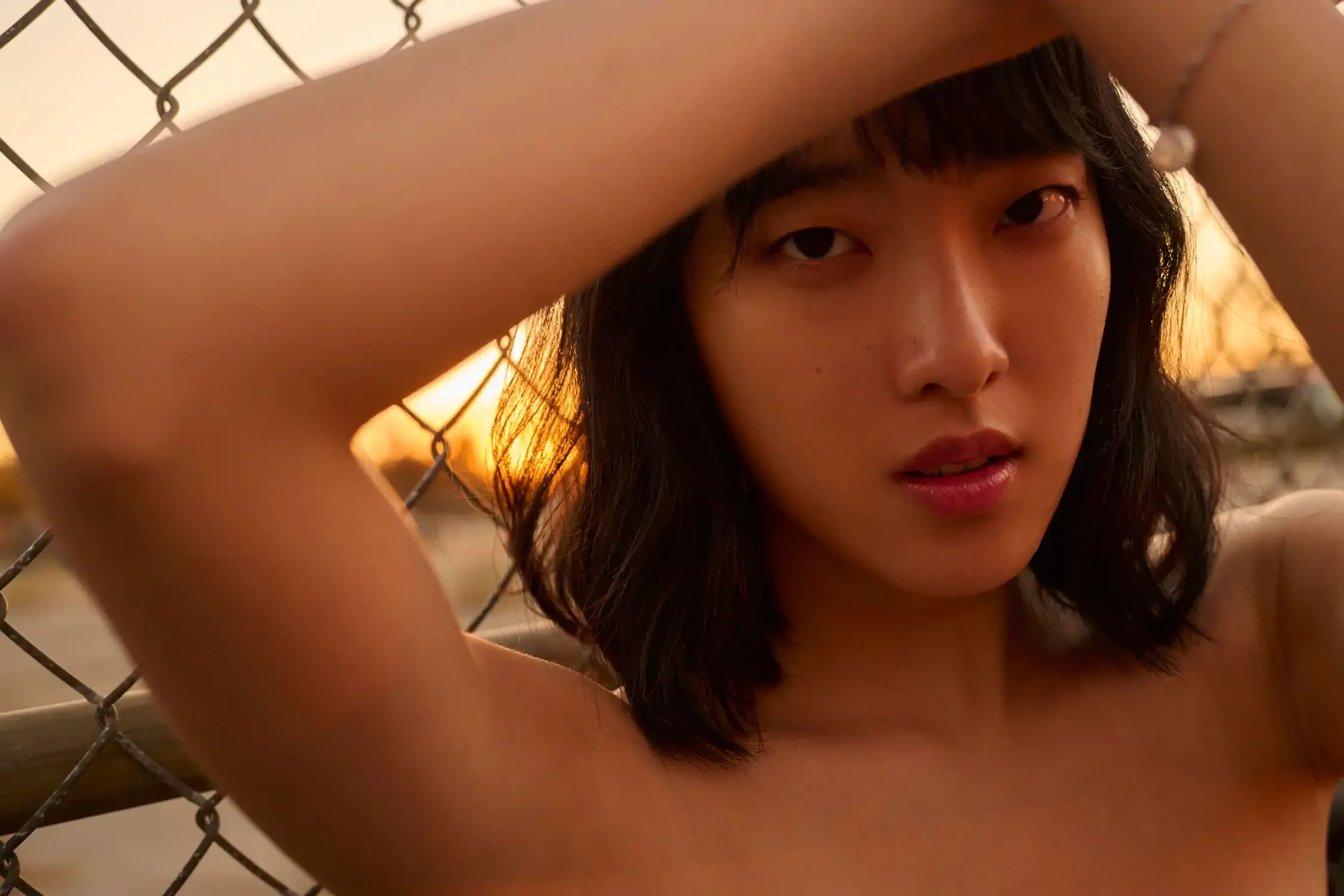 A young Asian woman with dark hair and brown eyes looking through a chain-link fence at sunset.