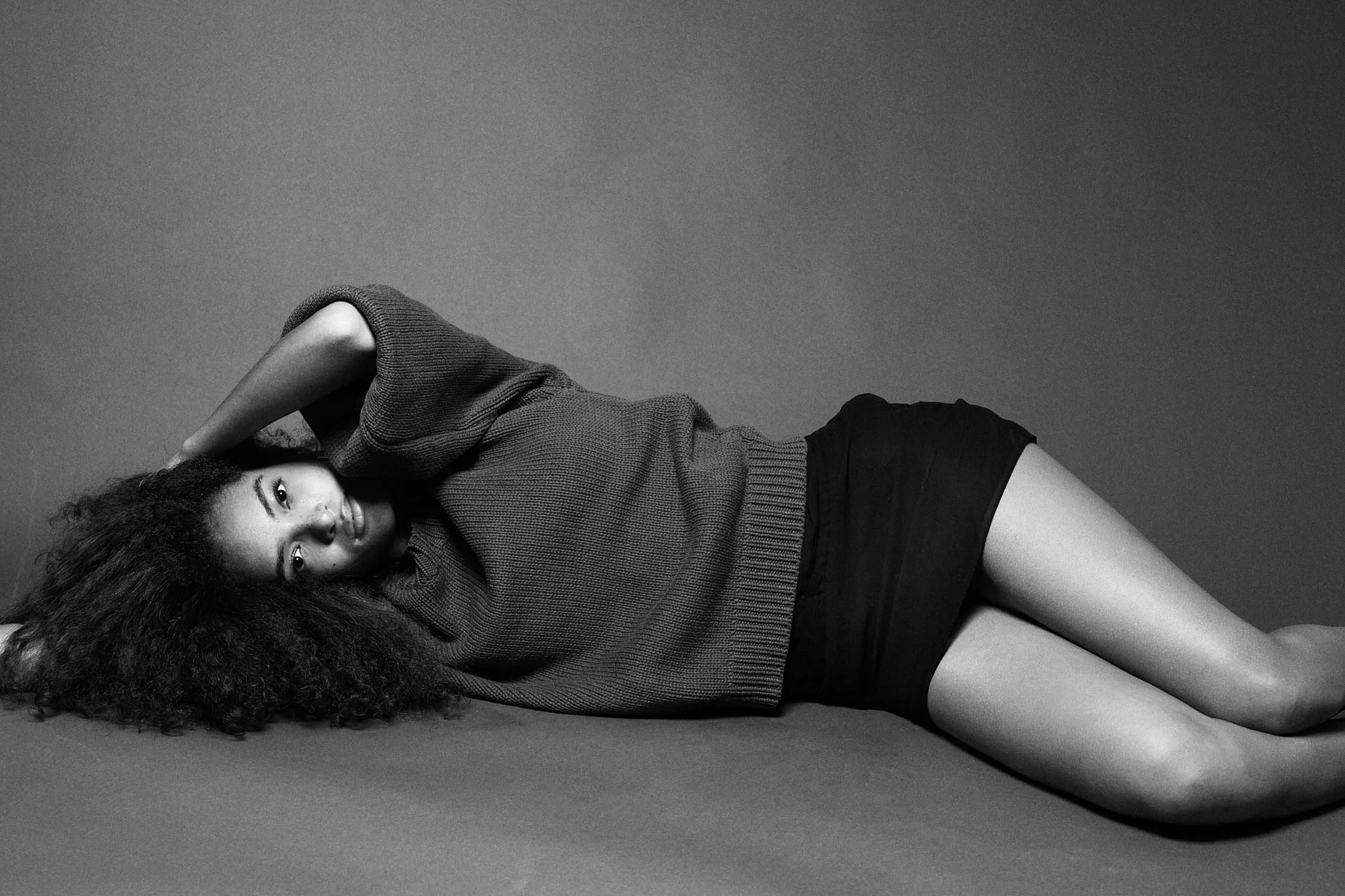 A black-and-white photo of a woman lying on her side on a plain surface, with her head resting on one arm and her legs slightly bent. She has curly hair, wears a loose sweater and shorts, and looks directly at the camera.