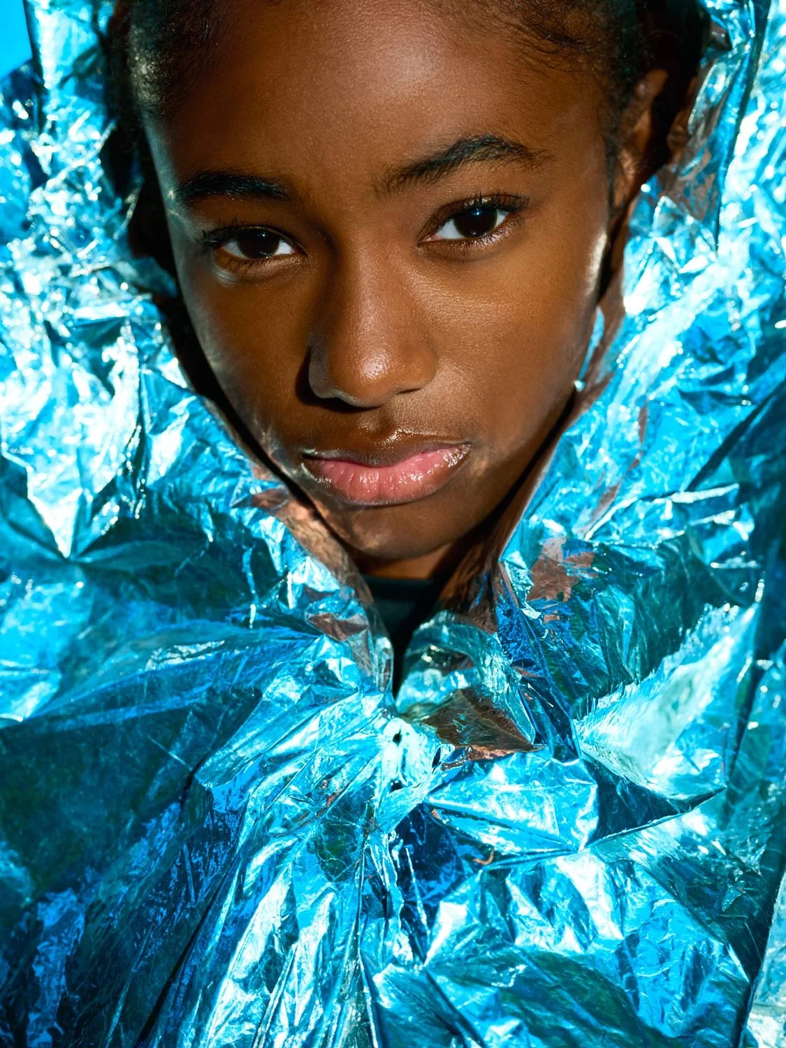 Close-up of a young woman with dark skin and dark hair, wrapped in shiny, metallic blue foil.
