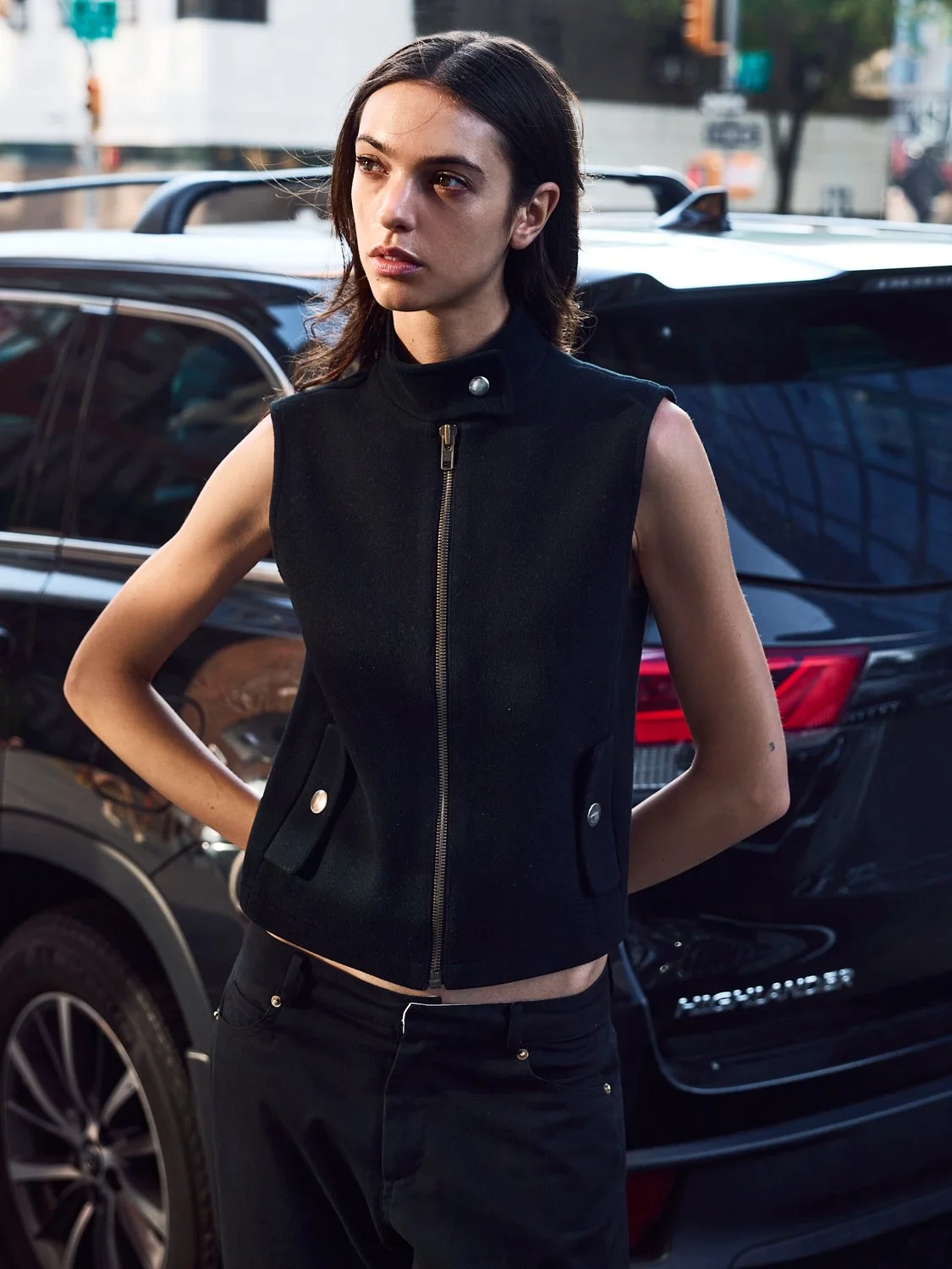 A woman standing outdoors in front of a black vehicle, wearing a sleeveless black vest with a gold zipper, and black pants.