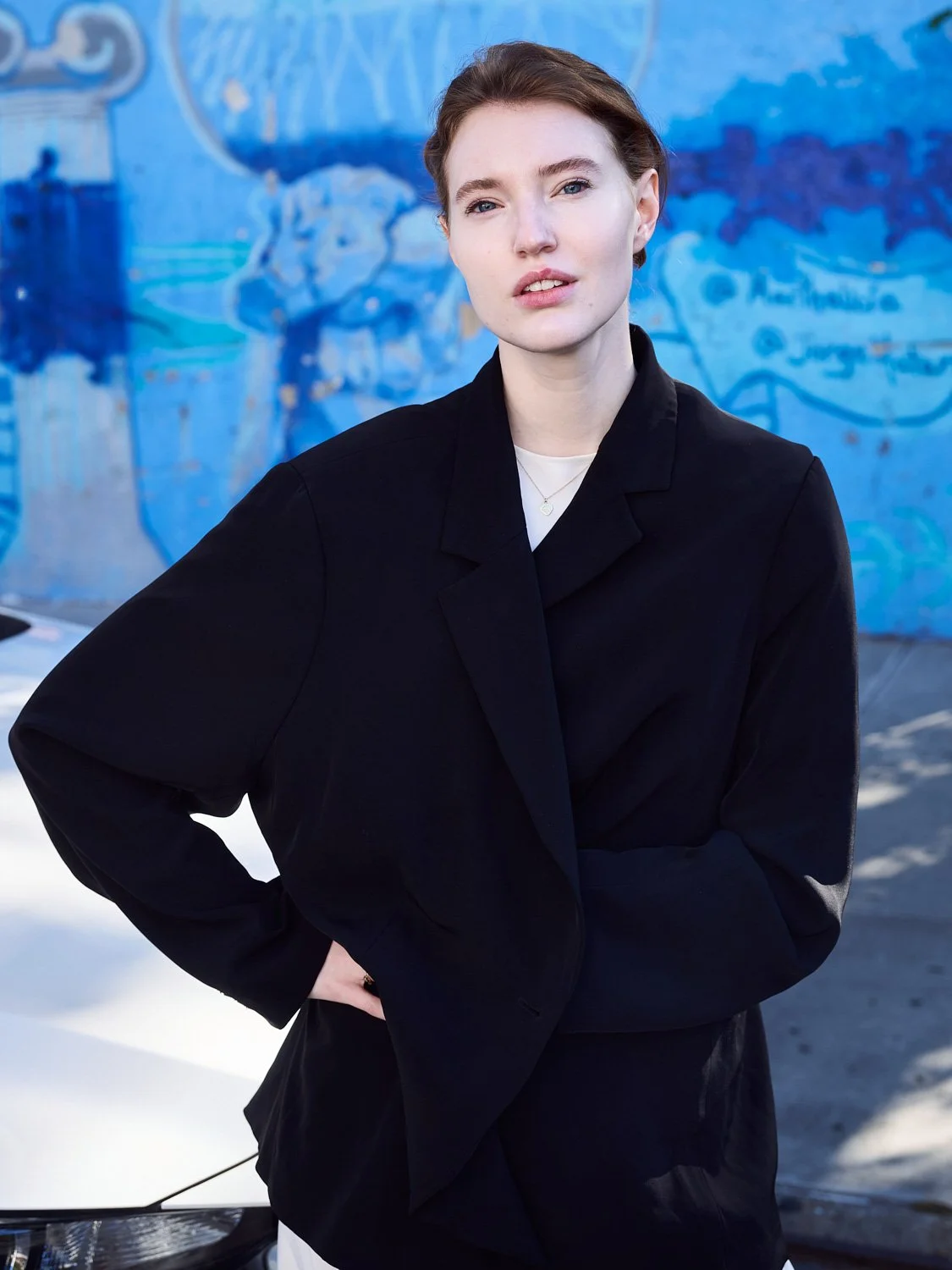 A woman with short brown hair and fair skin wearing a black blazer and white top, standing outdoors in front of a blue wall with a mural.