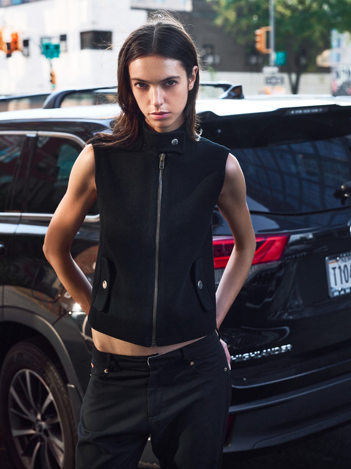 A young woman with dark brown hair and a serious expression standing outdoors in front of a black SUV car on a city street, wearing a sleeveless black vest and black pants.