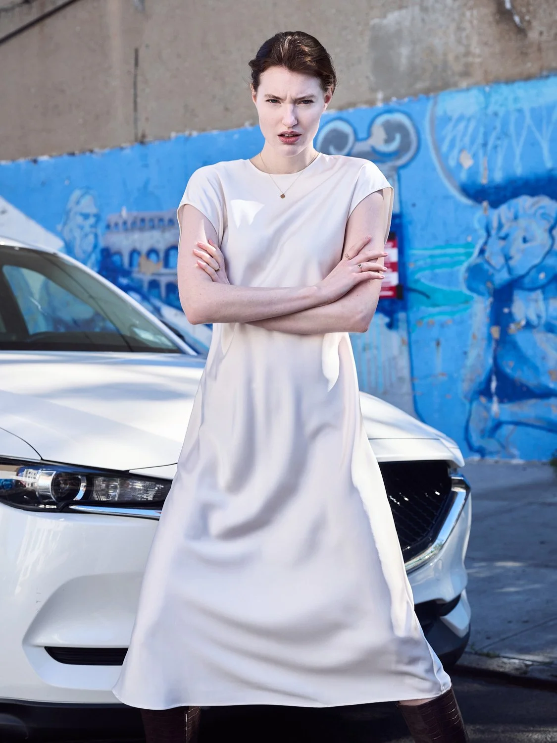 A woman in a white dress standing with arms crossed in front of a white car, with a blue mural painted on the wall behind her.