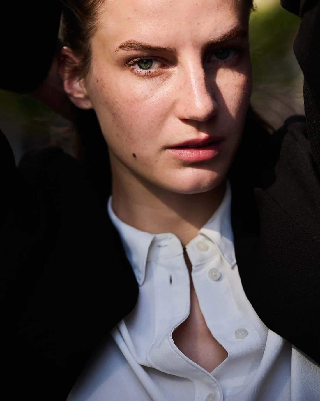 Close-up of a woman with fair skin and light green eyes, wearing a white shirt and black outerwear, looking intently at the camera with her arms raised behind her head.