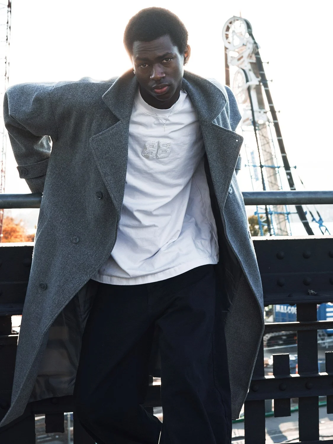 A young man leaning on a railing outdoors, wearing a gray coat over a white T-shirt with a graphic design, dark pants, and a silver cross necklace, with a Ferris wheel in the background.