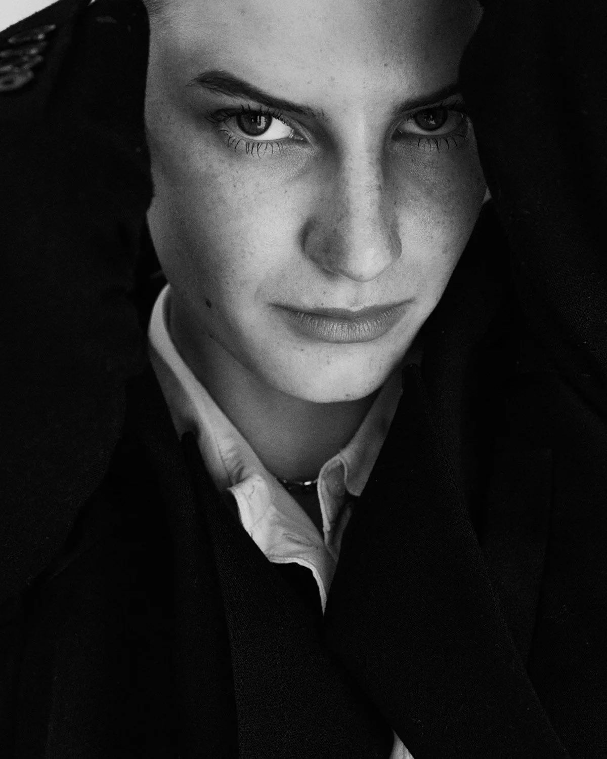 
Black and white beauty shot of a woman with fair skin and light eyes, wearing a white shirt and black outerwear, looking intently at the camera in. beautiful lighting.