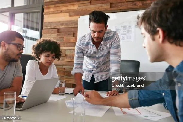 Four people in a meeting room collaborating around a table, with a laptop, papers, and drinks, in a modern office with wooden wall decor.