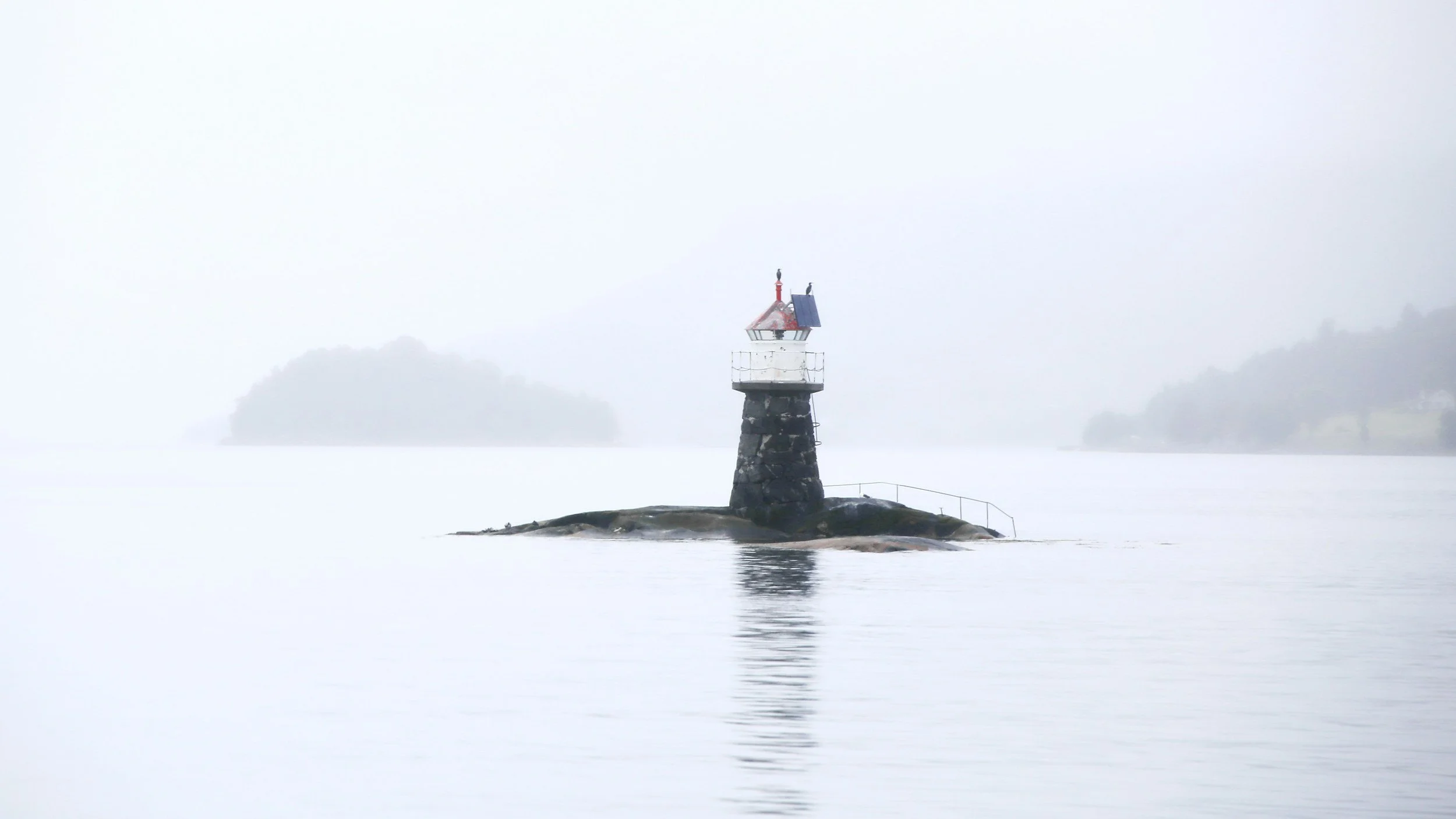 A solitary lighthouse on a rocky rock formation in the water, surrounded by misty, overcast sky and distant blurred trees.