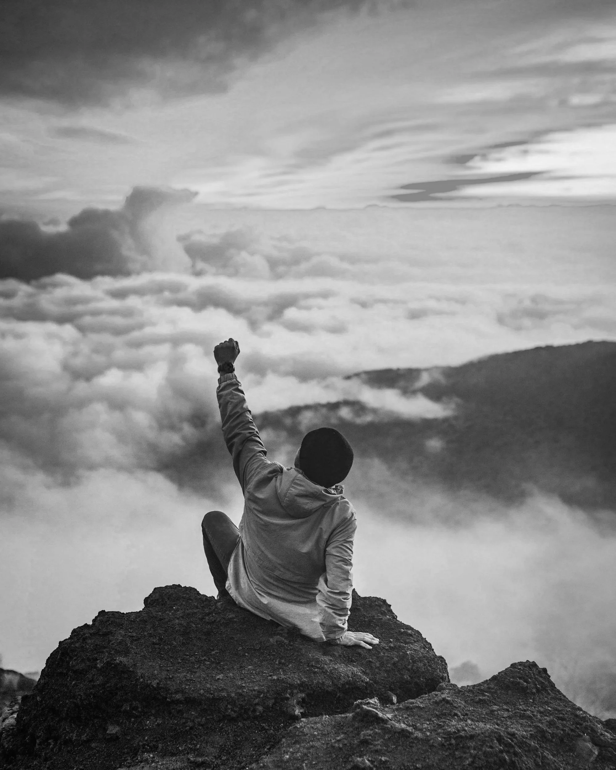 A person sitting on a rocky cliff overlooking a sea of clouds, raising a fist in victory or celebration, in black and white.