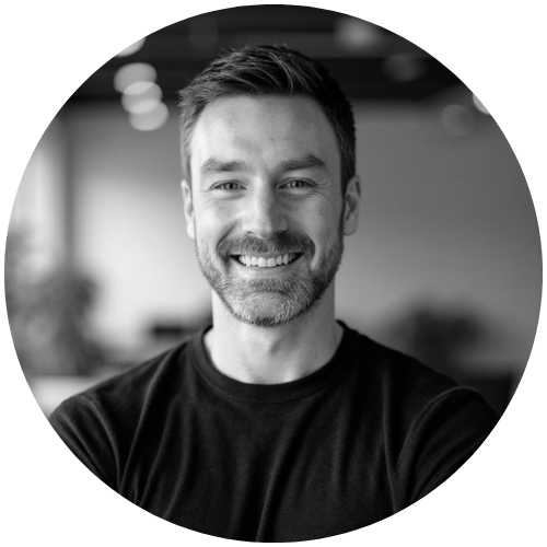 Black and white photo of a smiling man with short hair and a beard, wearing a black T-shirt, in an indoor setting.