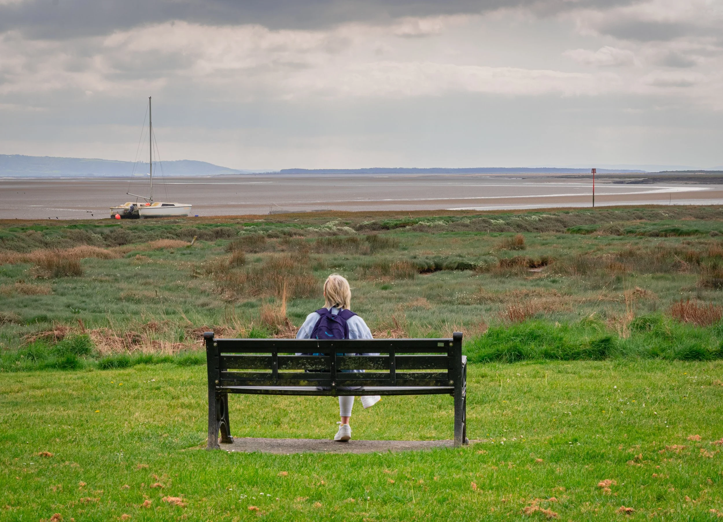 A person with blonde hair, wearing a blue jacket, white leggings, and sneakers, on a park bench overlooking a beach with a sailboat, water, and cloudy sky in the background.