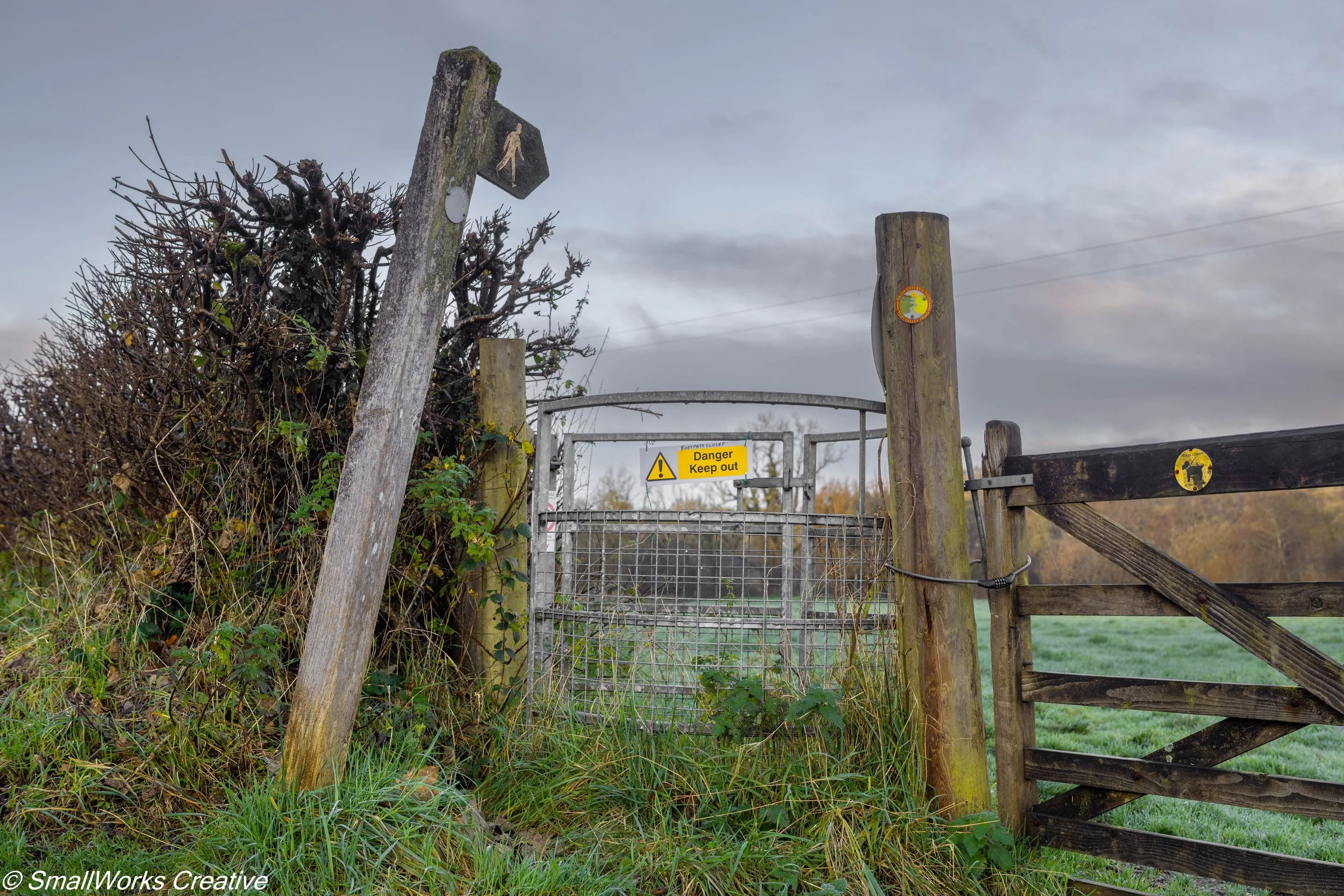 A gated rural pathway with a sign that says 'Danger Keep out', surrounded by overgrown grassy and bush areas, and a cloudy sky in the background.