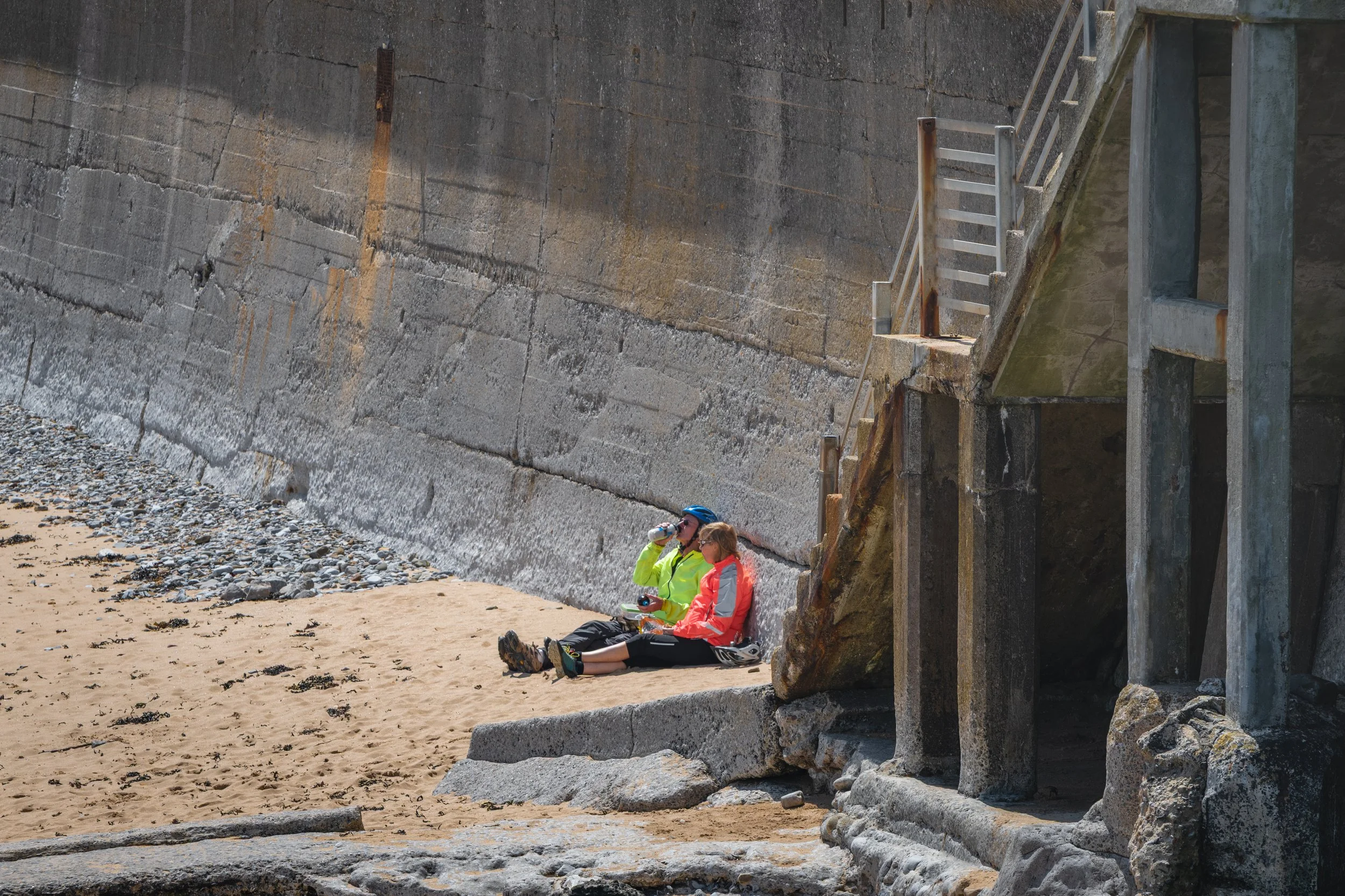 Two people sitting on the beach, drinking from bottles while leaning against a concrete wall with stairs and a small building structure nearby.