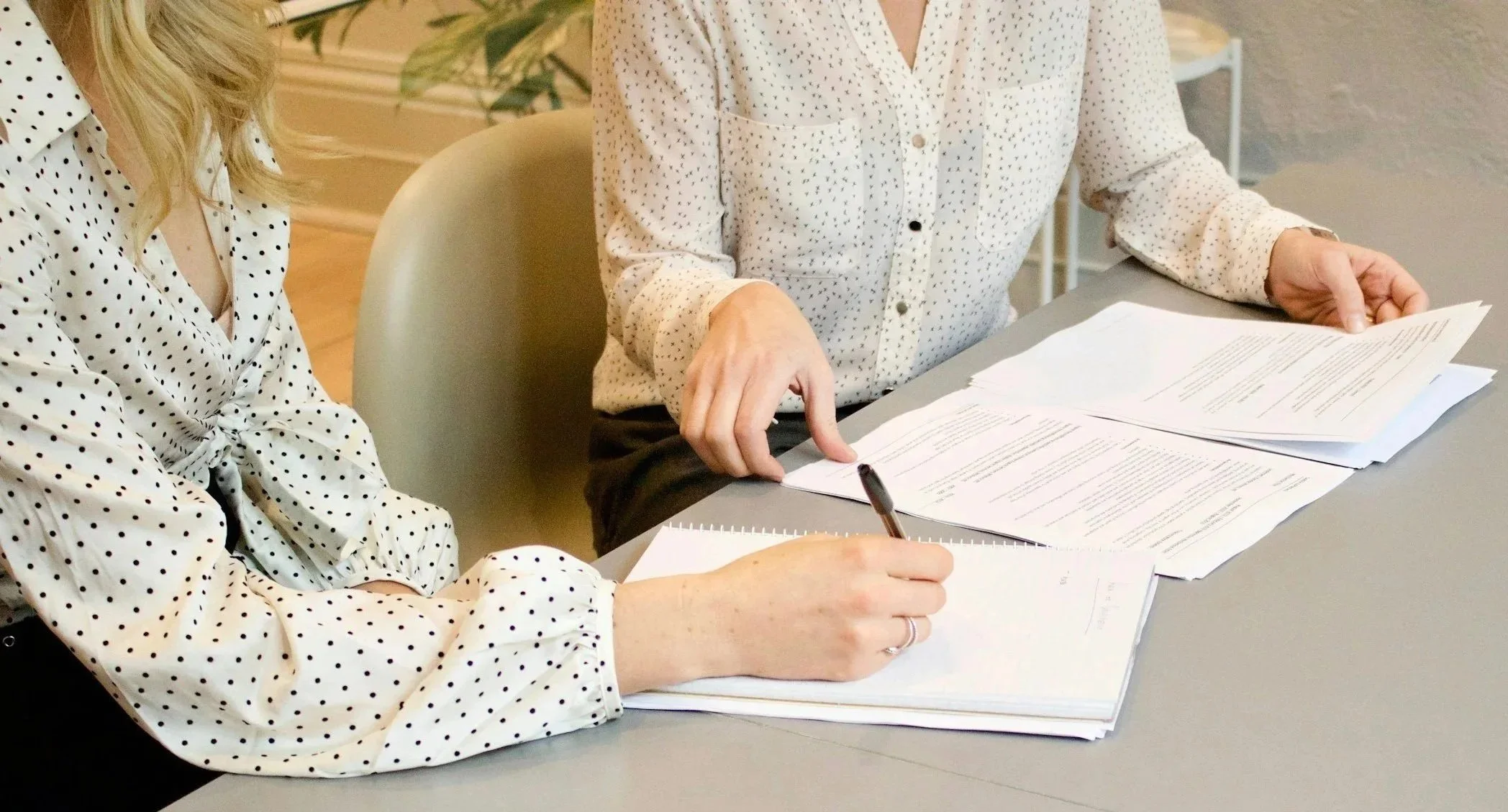 Two women in polka dot blouses sit at a table reviewing papers, with one taking notes with a pen.