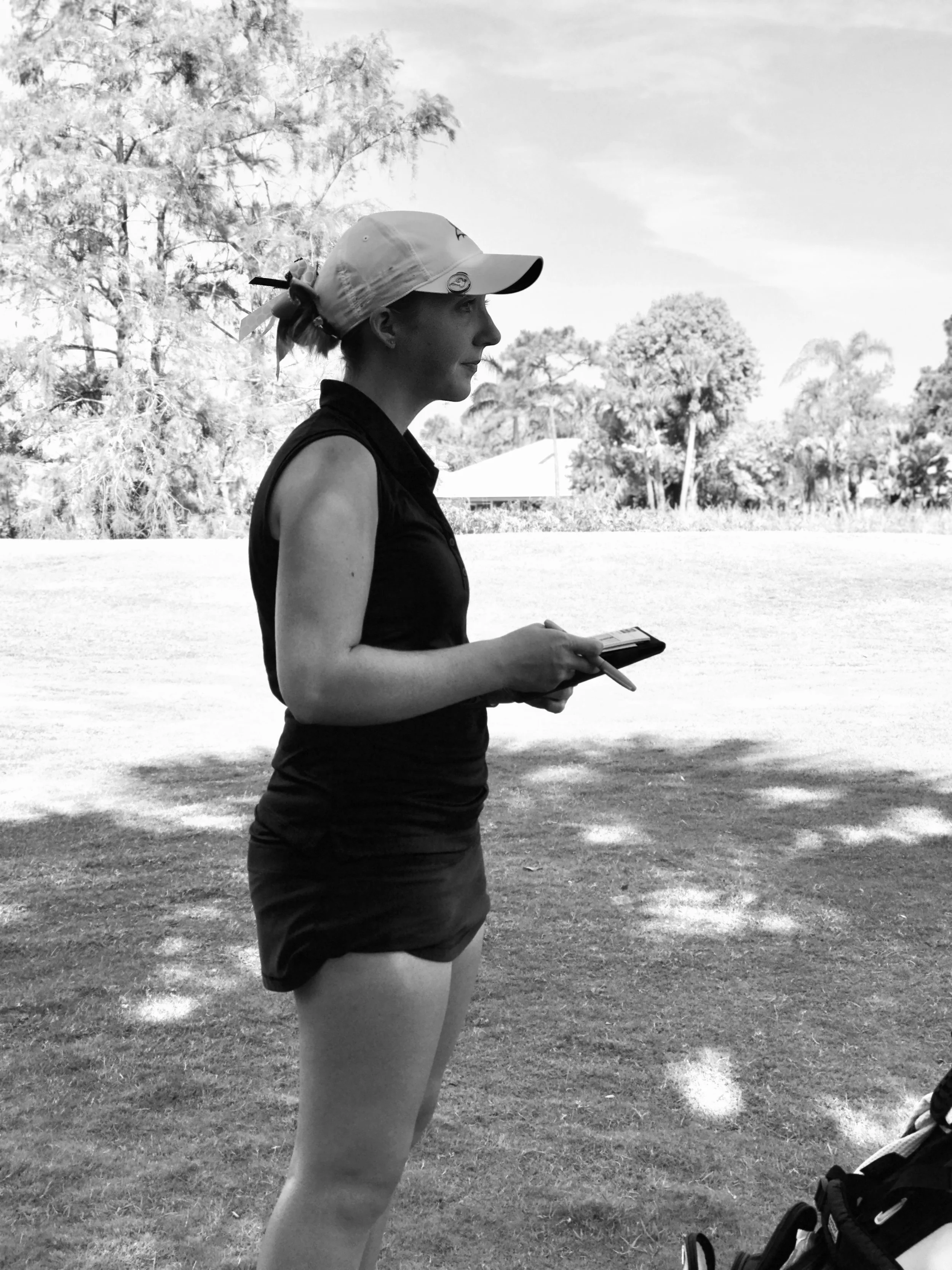 A woman wearing a hat and athletic outfit holding a scorebook outdoors at a golf course with trees in the background.