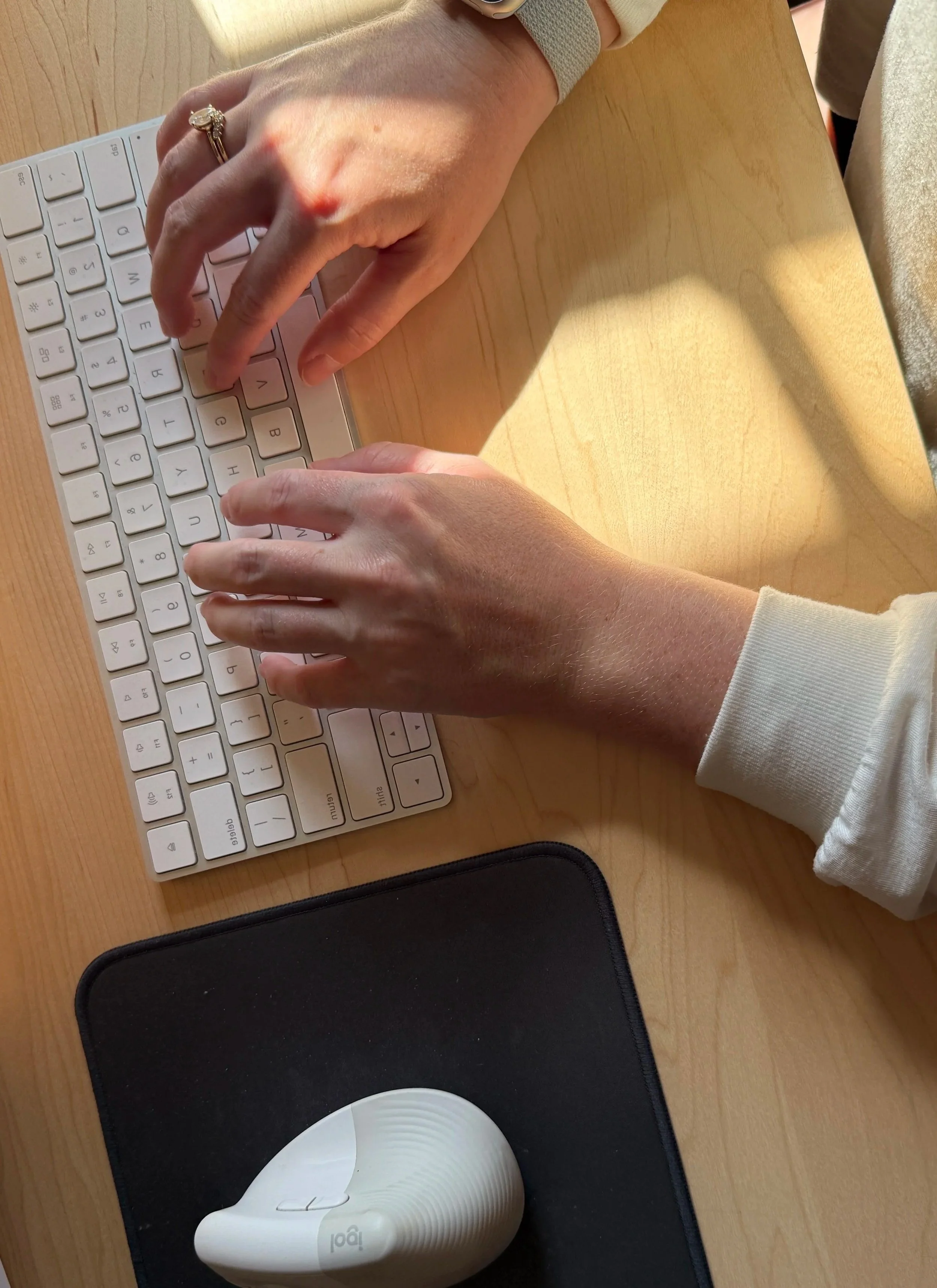 A person typing on a white keyboard at a wooden desk, with a black mouse pad, a black phone case, and a white disposable coffee cup nearby.