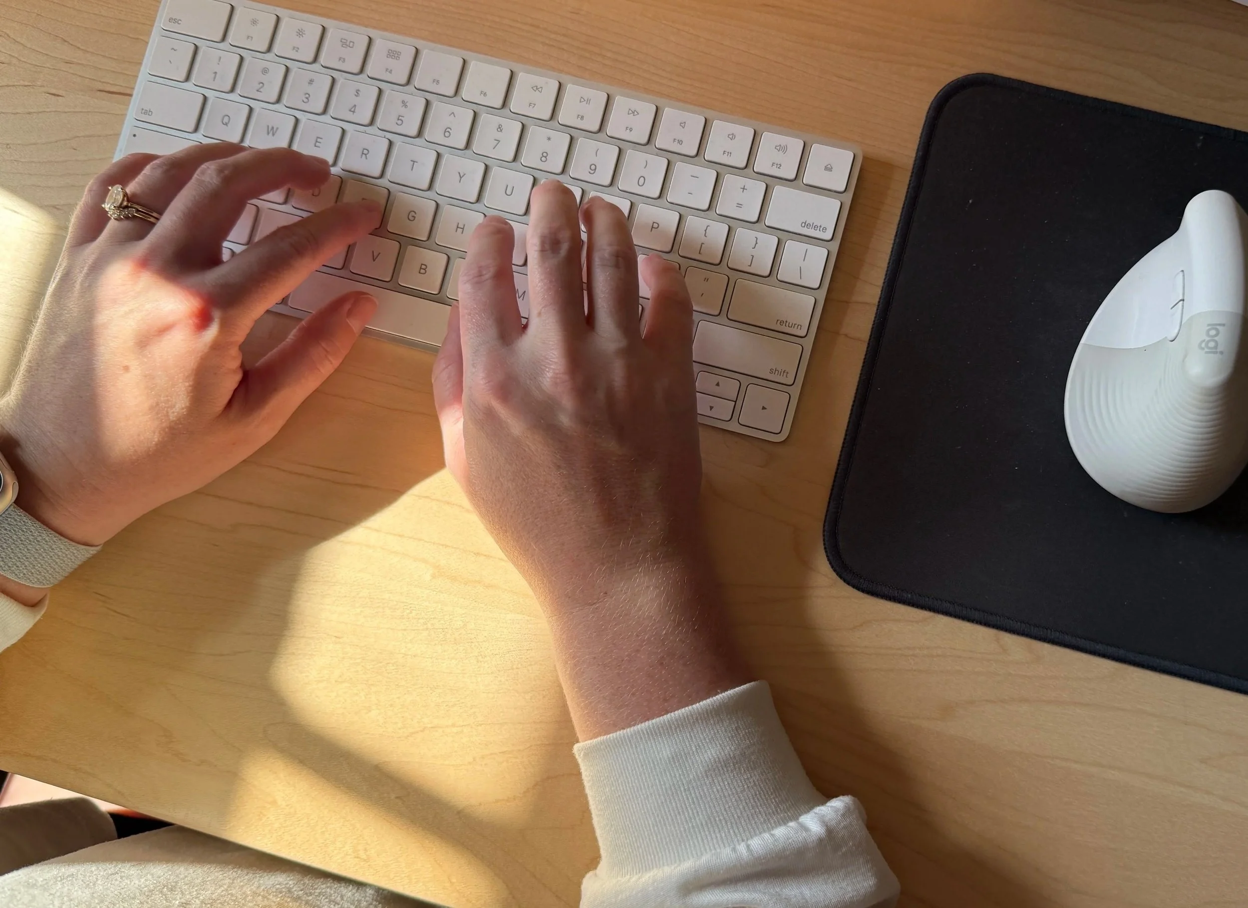 A person typing on a white wireless keyboard on a light wooden desk, with a black mouse pad and an ergonomic mouse nearby.