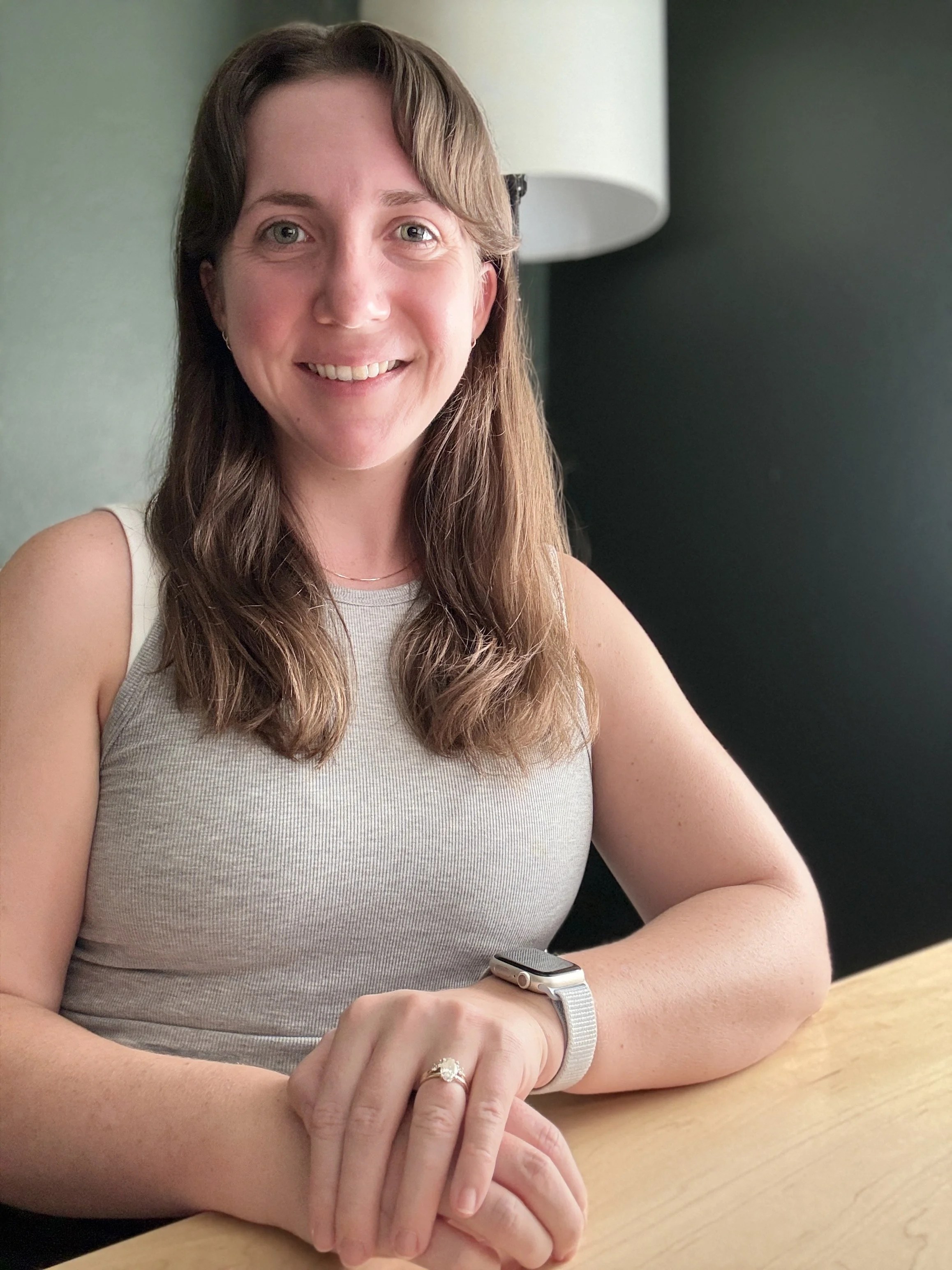A woman with long brown hair, wearing a grey sleeveless top, smiling while sitting at a wooden table, with a smartwatch on her left wrist, a ring on her left hand, and a background featuring a lamp and a dark wall.