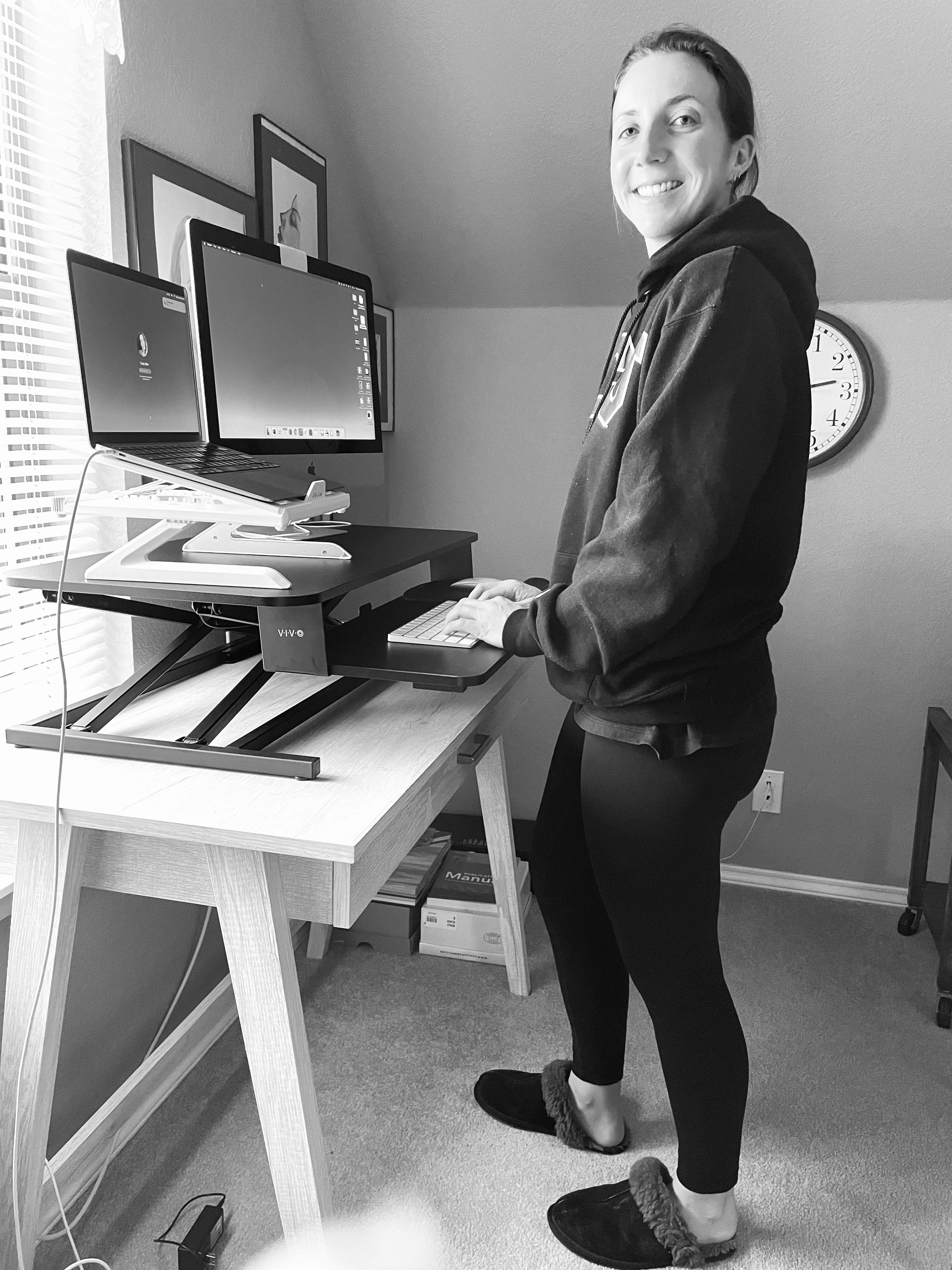 A woman standing at a standing desk with two monitors, smiling, in a room with framed pictures on the wall and a clock showing approximately 4:00.