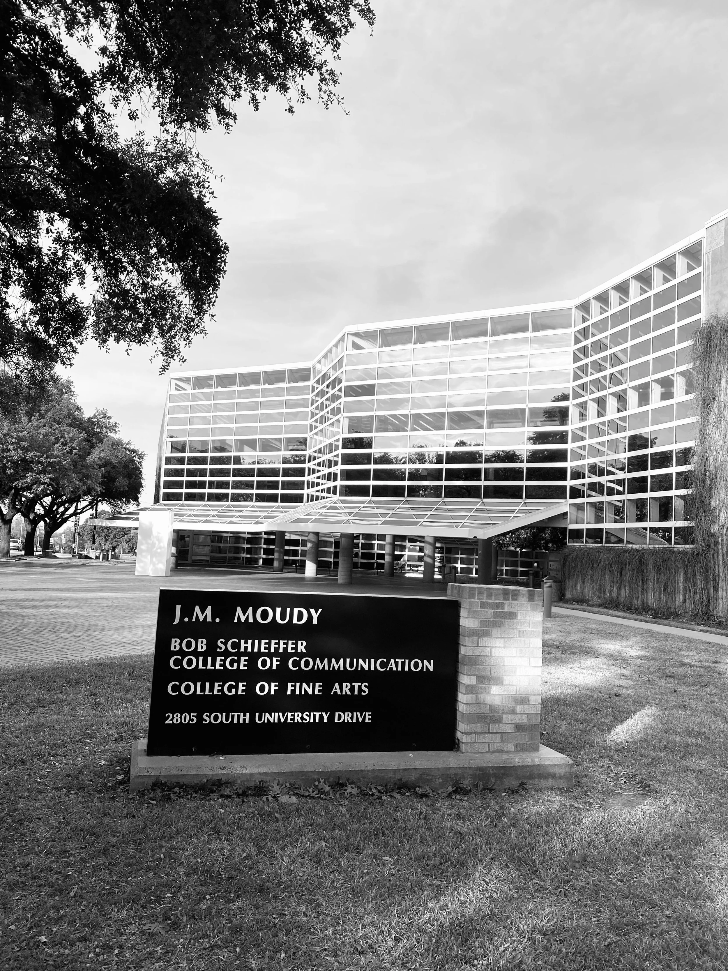 Photo of a modern multi-story building with glass windows, trees in the surroundings, and a sign in the foreground indicating the location as J.M. Moudy, Bob Schieffer College of Communication, College of Fine Arts at 2805 South University Drive.