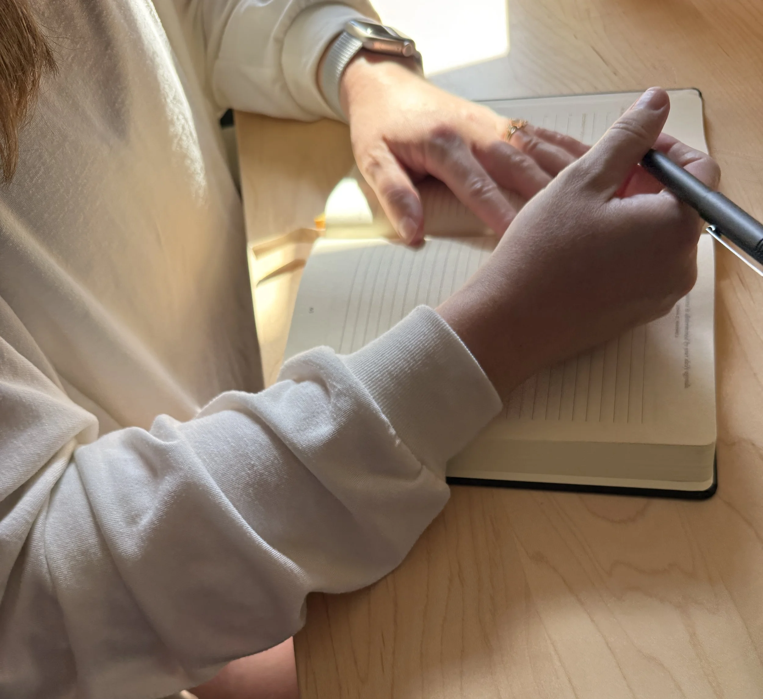 A person wearing a beige hoodie and a silver wristwatch is writing in a lined notebook with a black pen on a wooden table.