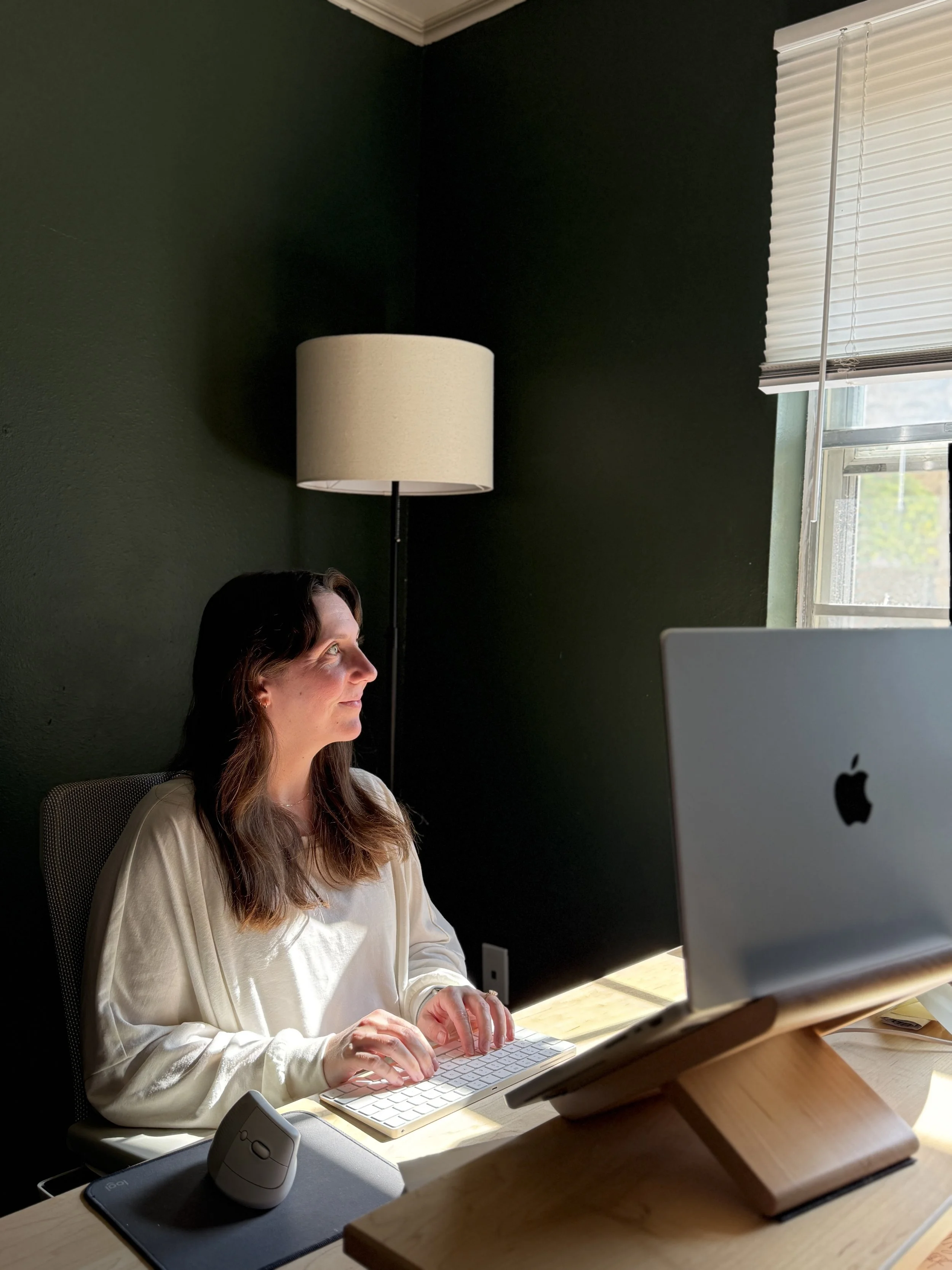 A woman with long brown hair in a white blouse working at a desk on a computer. There is a lamp behind her and a window with blinds on the right side.