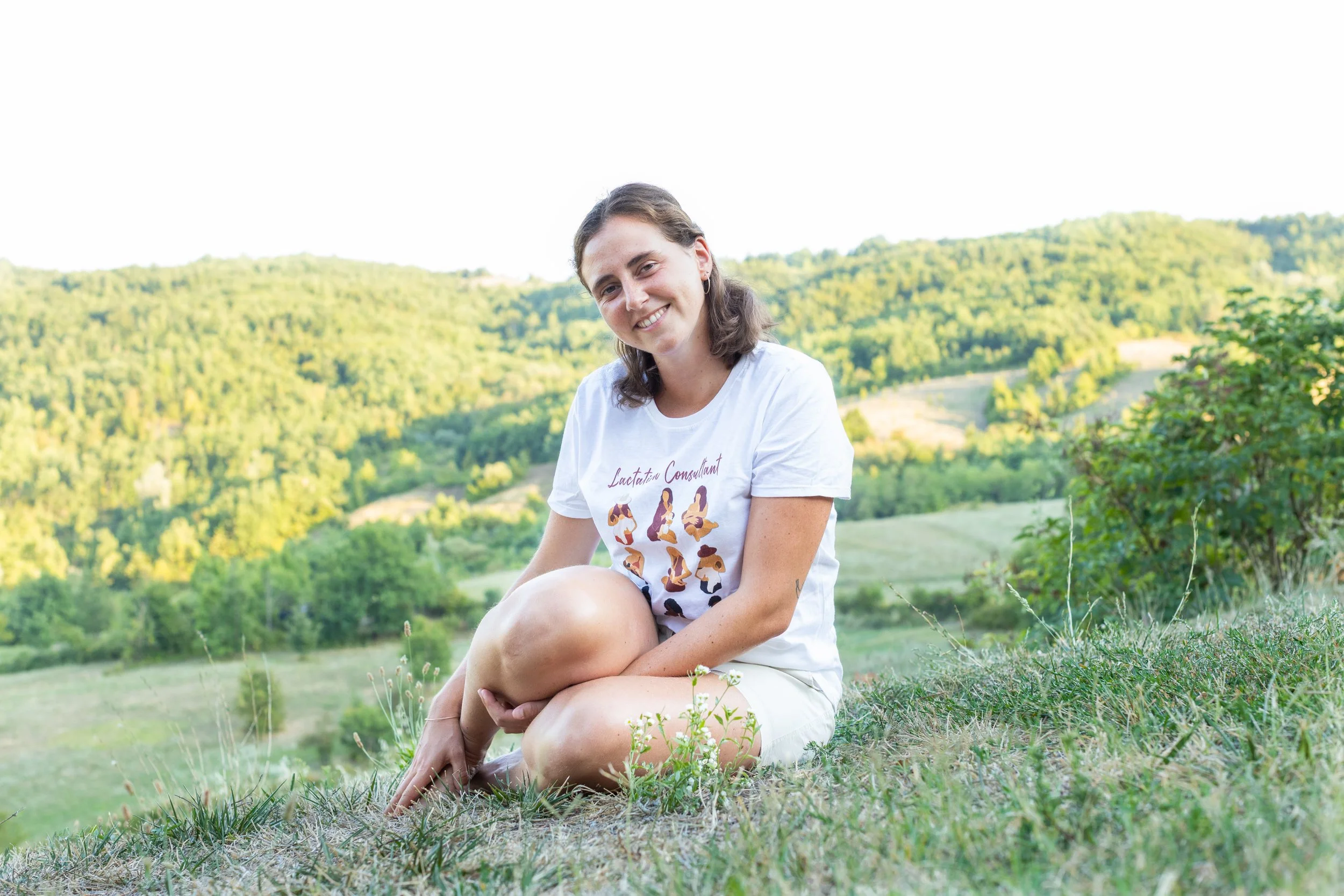 A woman sitting outdoors on grass in a scenic landscape of green hills and trees, smiling at the camera.