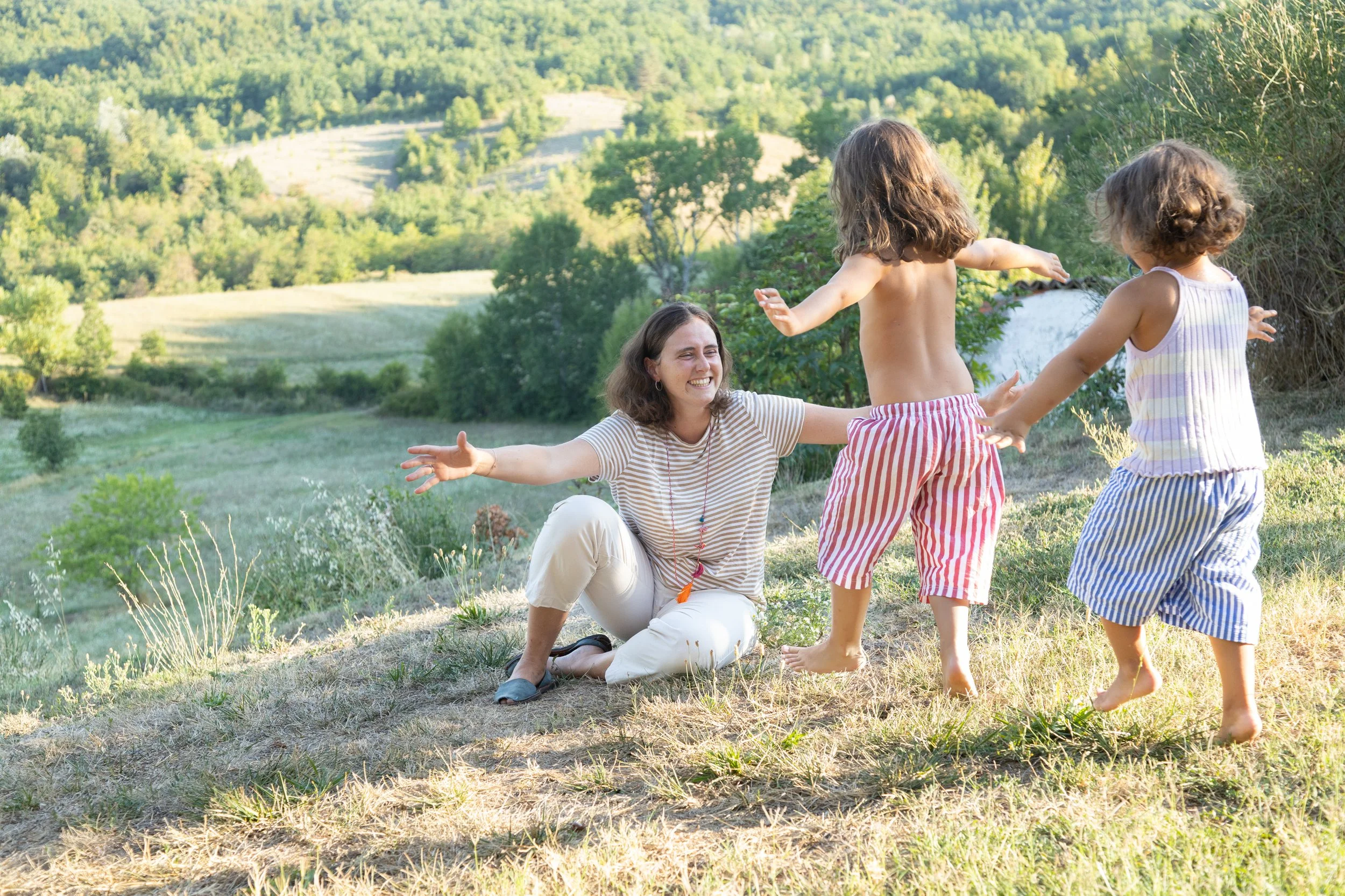 A woman playing and smiling with two children on a grassy hill in a lush, green landscape.